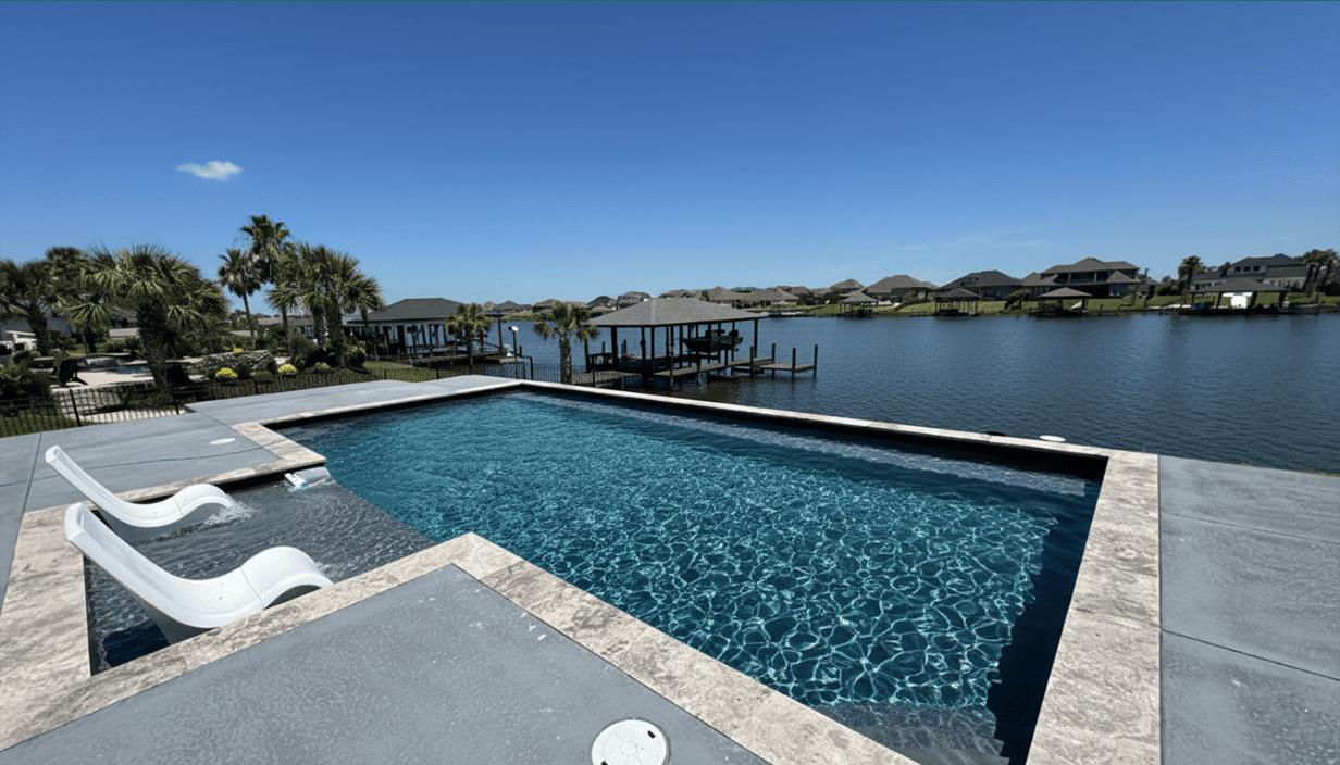 View of a swimming pool beside a waterway with boats, houses, and palm trees in the background under a clear blue sky.