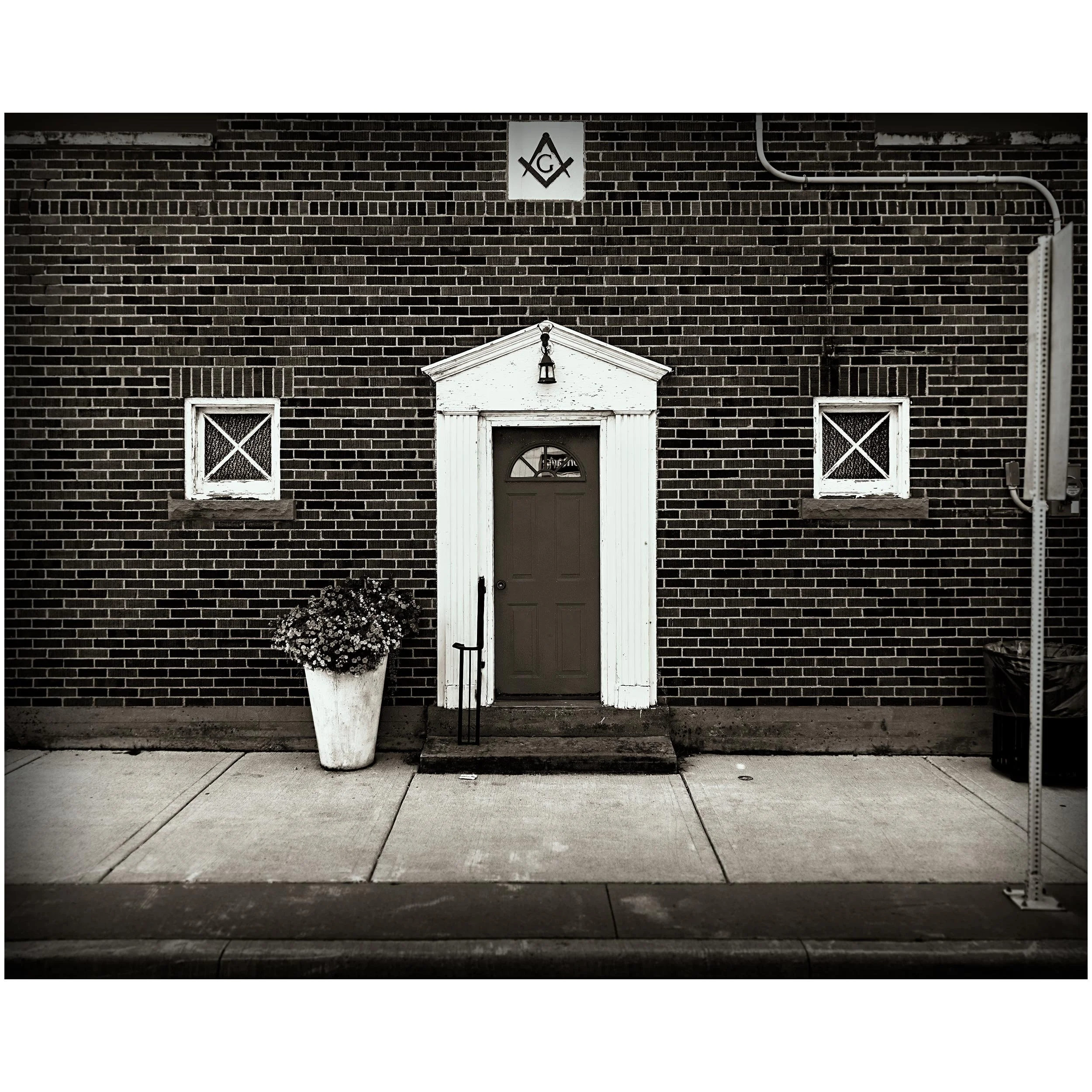 Black and white photo of a brick building with a porch, two small windows with X-shaped panes, a door with a window, a potted plant, and a sign with a square and a triangle above the door.