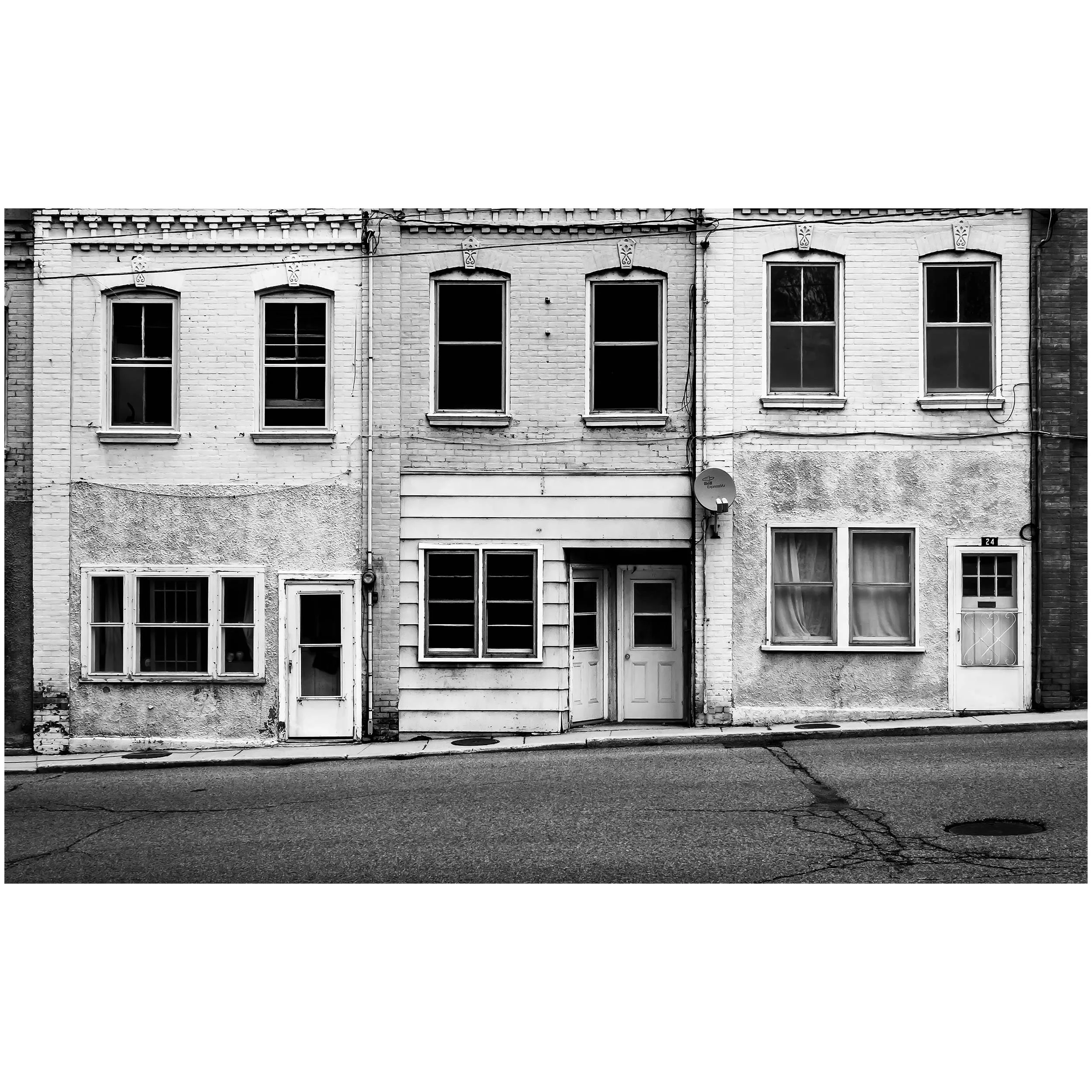 Black and white photo of an old brick row house with four windows on the upper floor, some with curtains, and a door on the ground floor. Utility wires run across the facade.