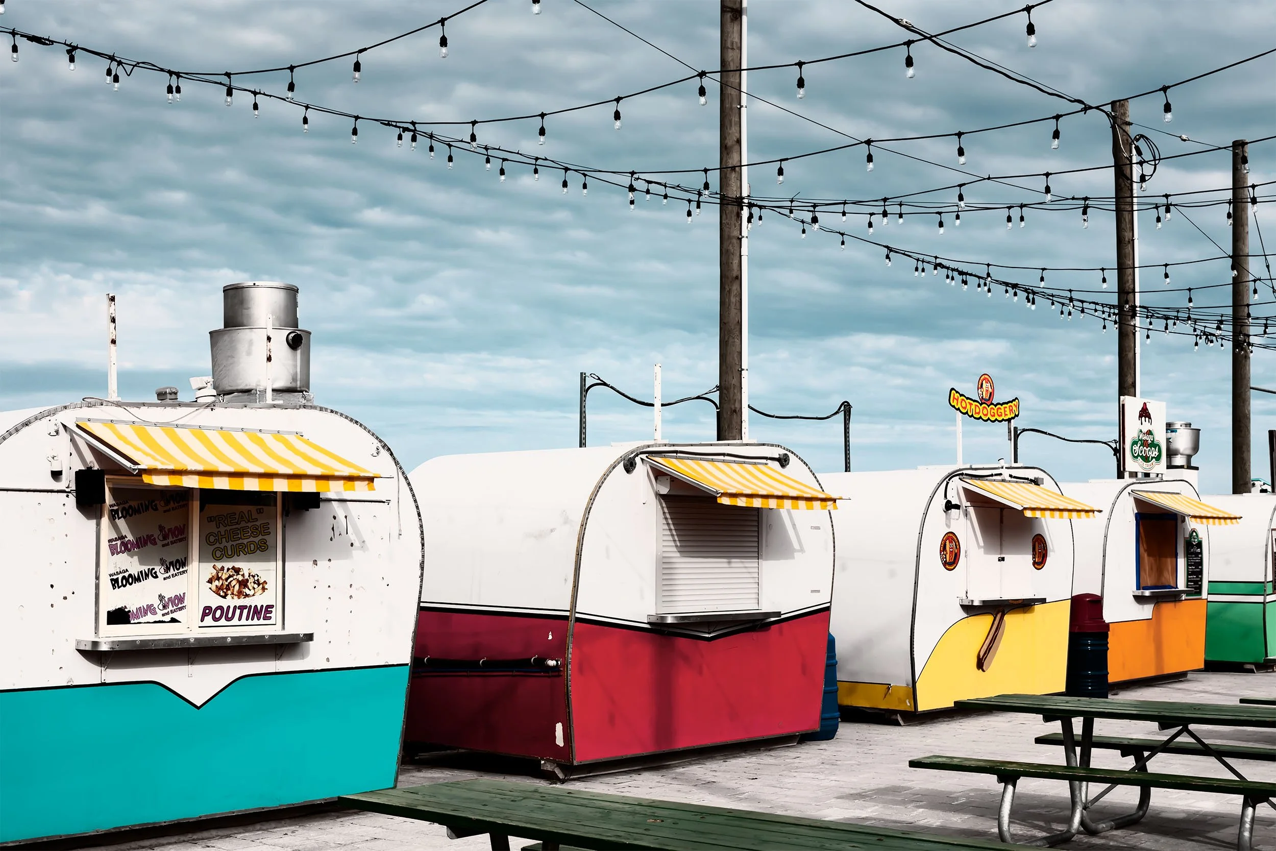 Colorful food trucks with yellow and white striped awnings, a sign for poutine, and string lights overhead at an outdoor fair or market against a cloudy sky.