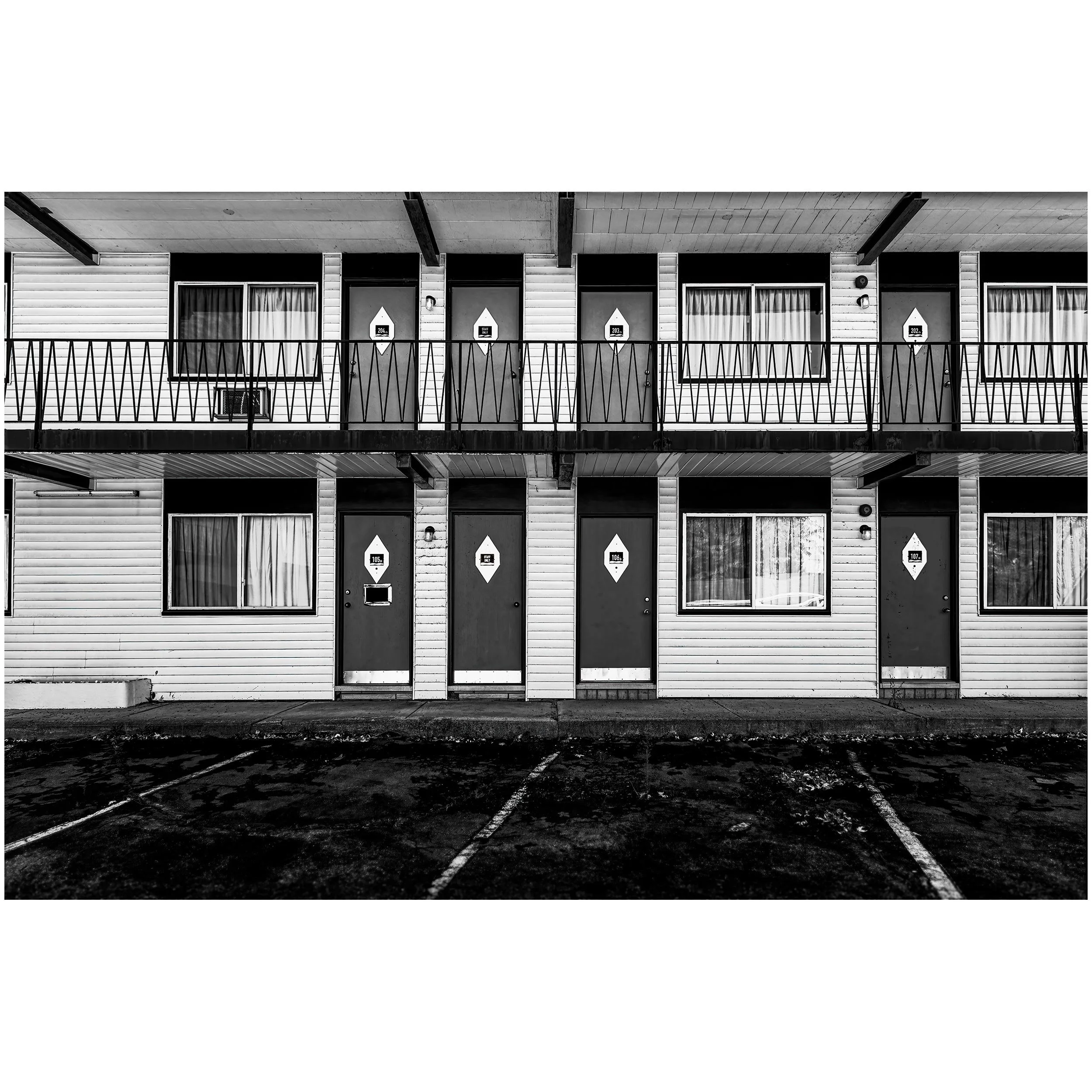Black and white photograph of an apartment building with four doors, a balcony, and a parking lot in the foreground.