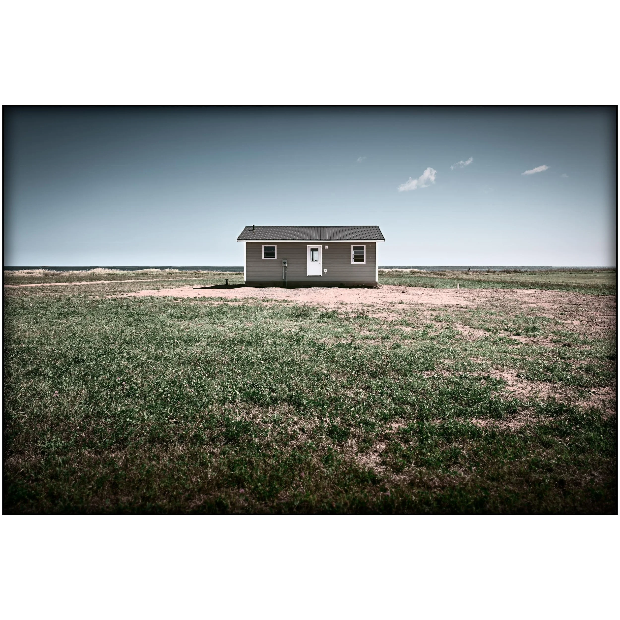simple grey building with a white door sits alone in a sparse grassy field near the shoreline under a wide pale sky.