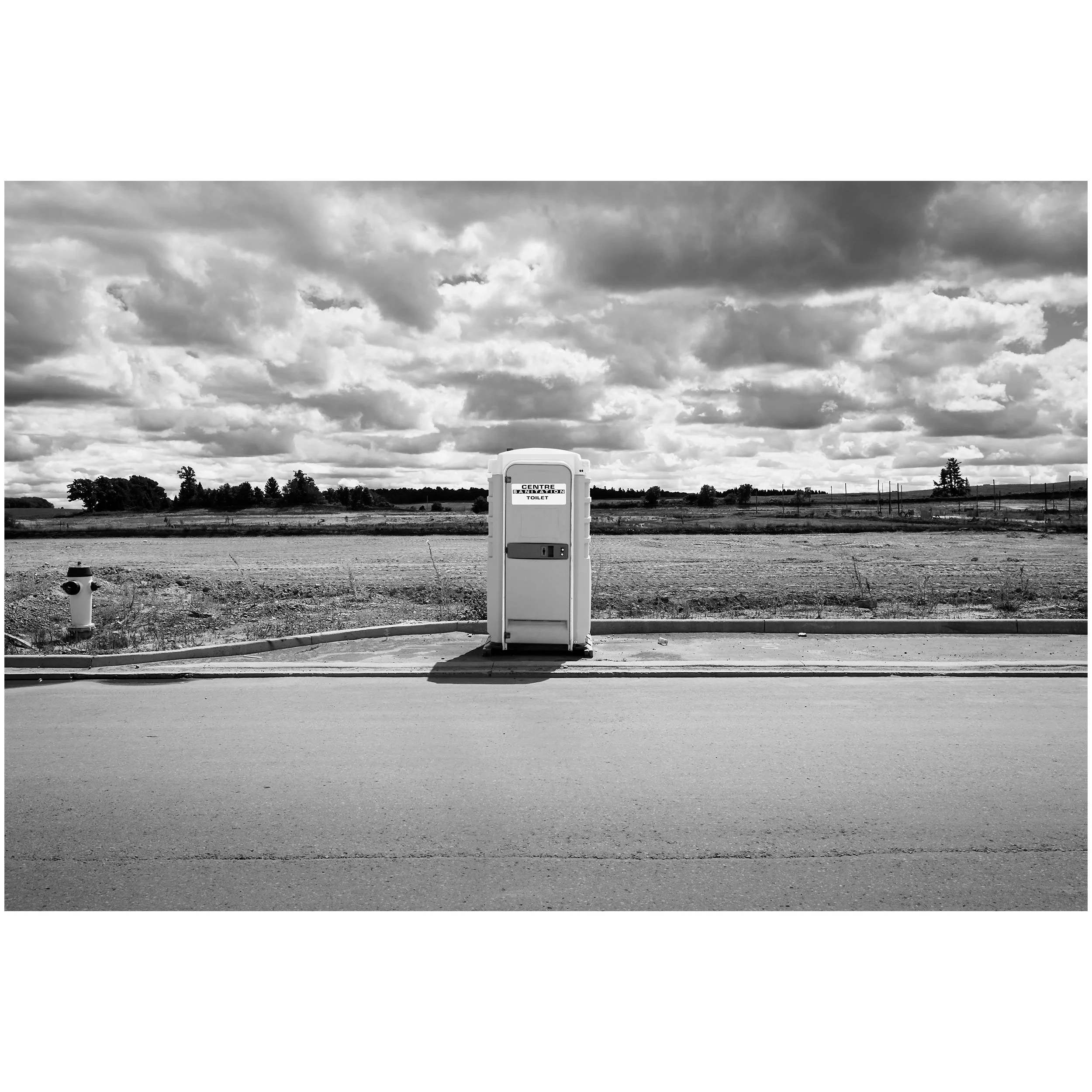 A black and white photograph of an open landscape with a cloudy sky, a portable toilet labeled "Centre sanitation toilet," and a fire hydrant in the foreground.