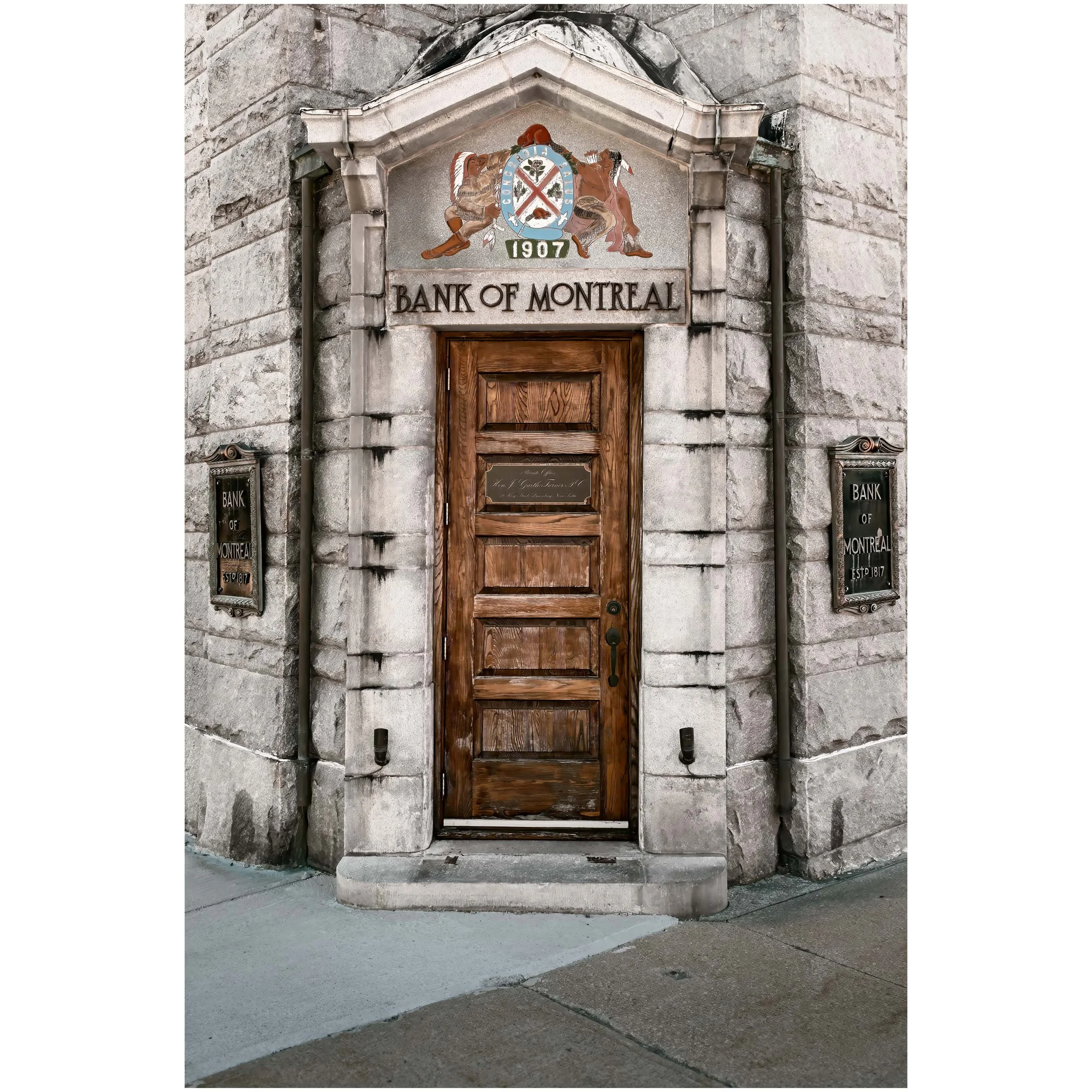 Stone building entrance with a wooden door, framed by a stone archway. Above the door is a colorful emblem featuring two figures, a shield, and the words 'Bank of Montreal' with the year 1907.