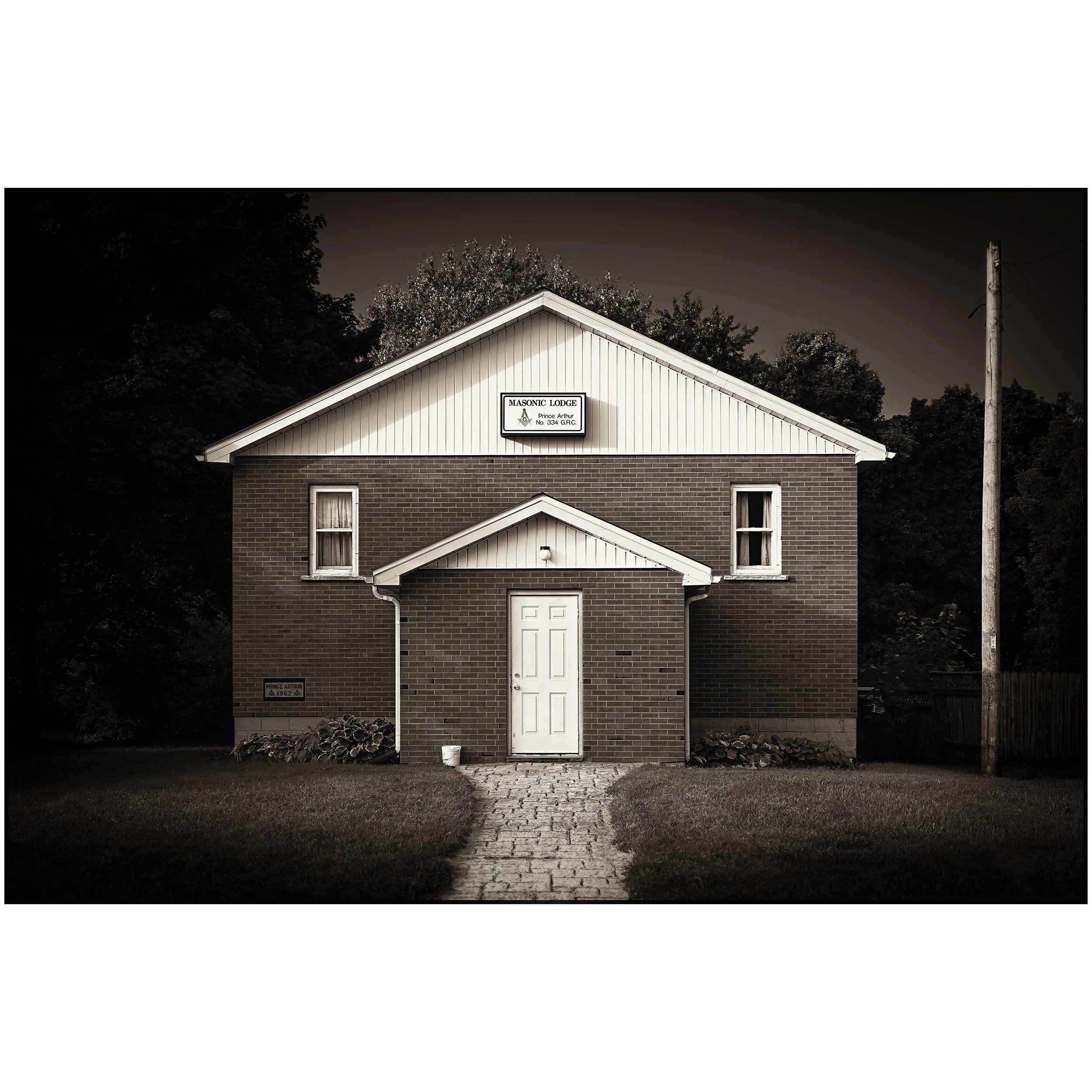 Black and white image of a brick church with a gabled roof, a sign that reads 'Masonic Lodge' and 'Prince Arthur No. 334 G.R.C.', a small front porch, and a stone-paved walkway leading to the door; there is a telephone pole on the right and trees in the background.