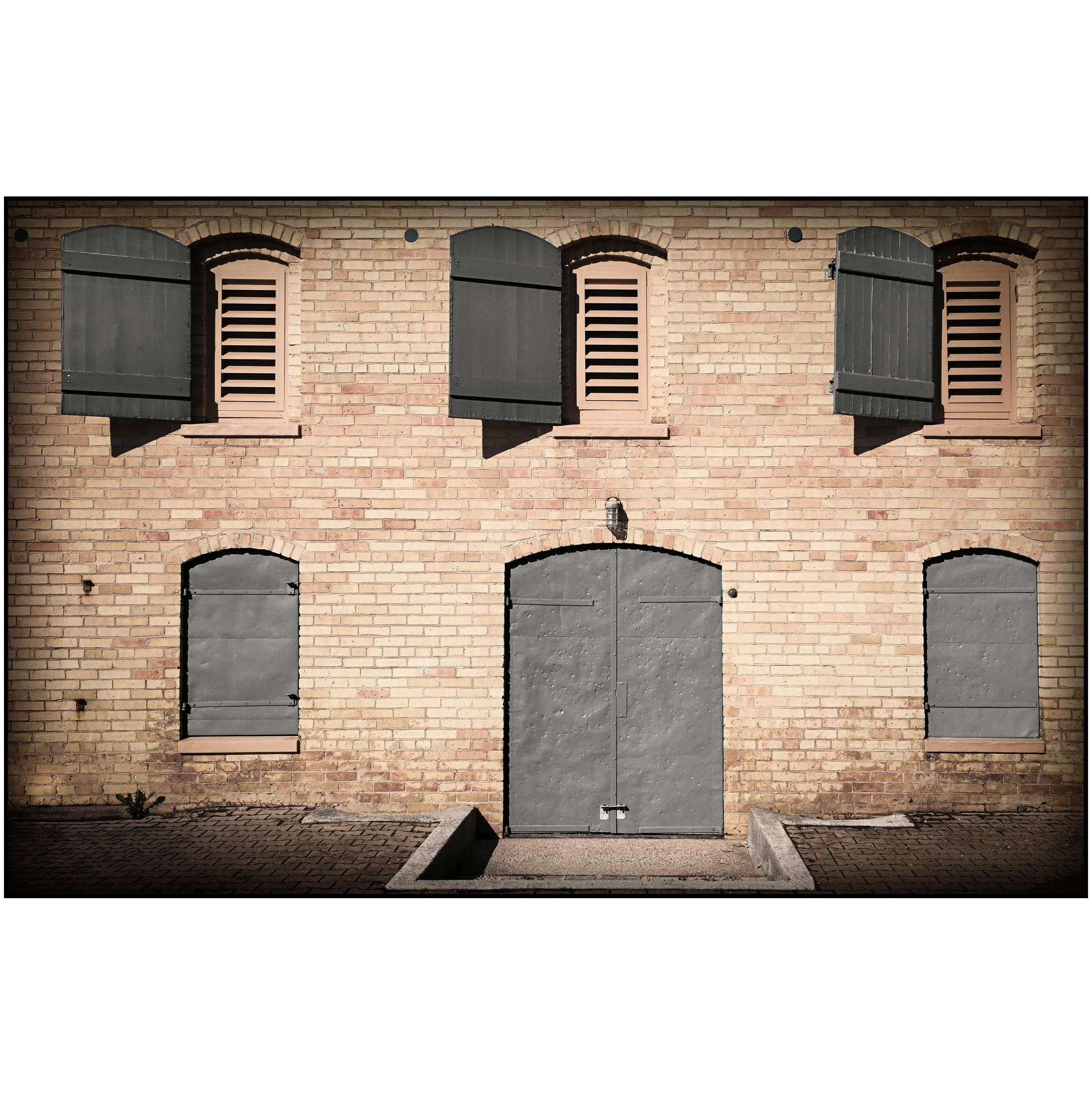 A brick building with three gray closed shutters and two black locks, three arched windows at the bottom, a smaller arched window at the top, and a small outdoor light fixture.