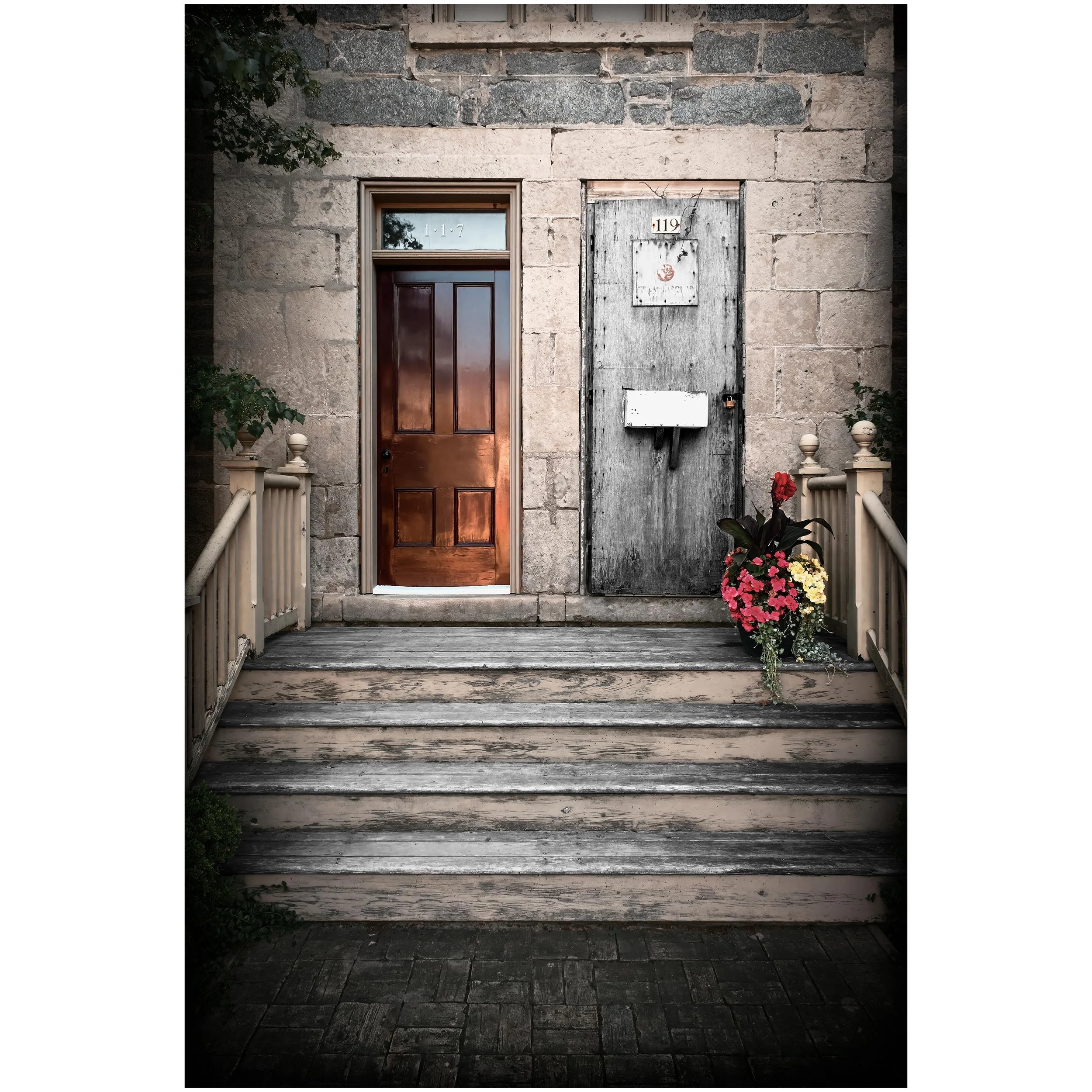 Exterior of a stone building with a wooden door numbered 119 and a window above it with the number 117. To the right of the door, there is a weathered metal mailbox and a flower pot with pink and white flowers. A white fence with decorative posts is in front of the entrance, and a small plant is visible on the left side.