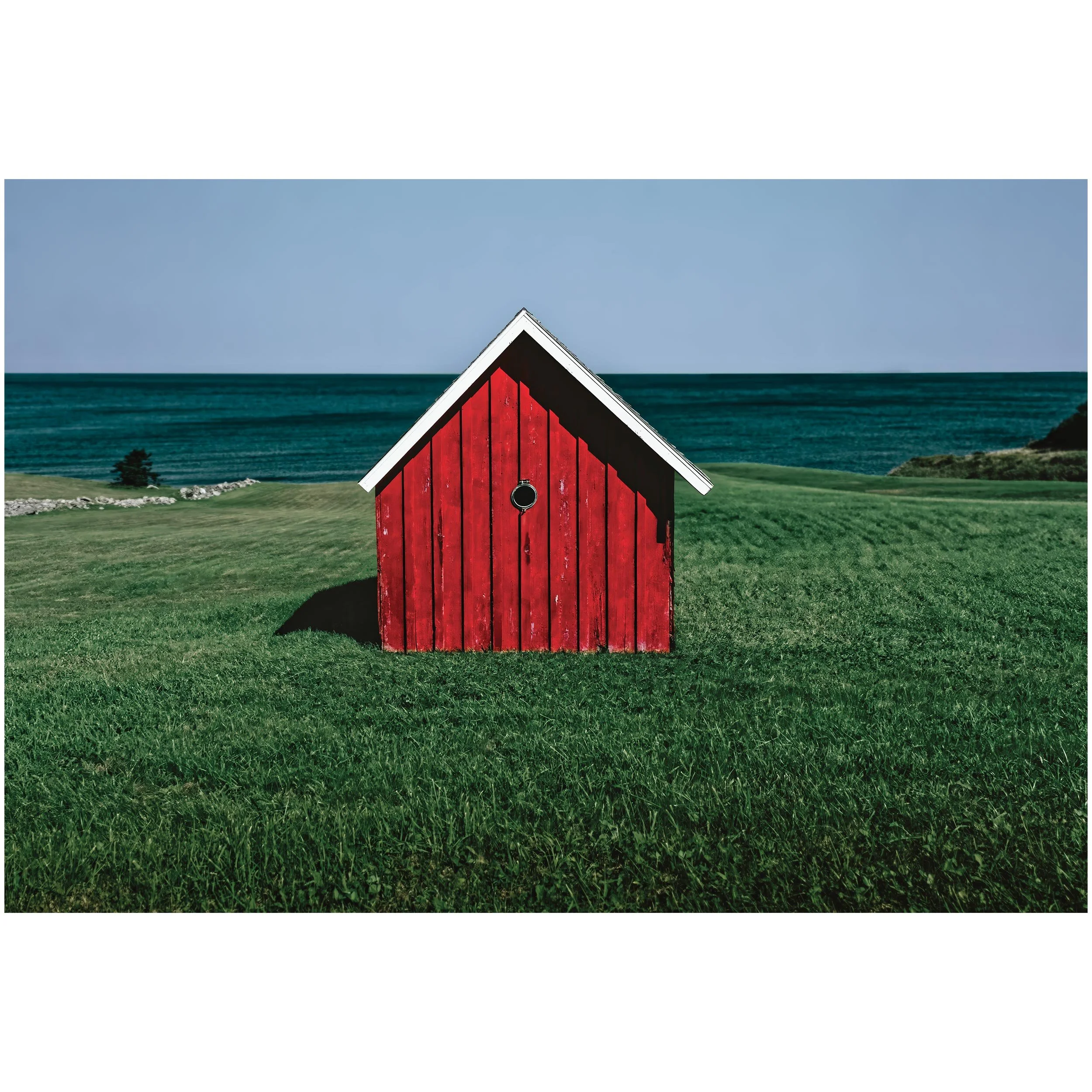 A red barn with white trim and black accents on a grassy field near a body of water, with a clear blue sky.