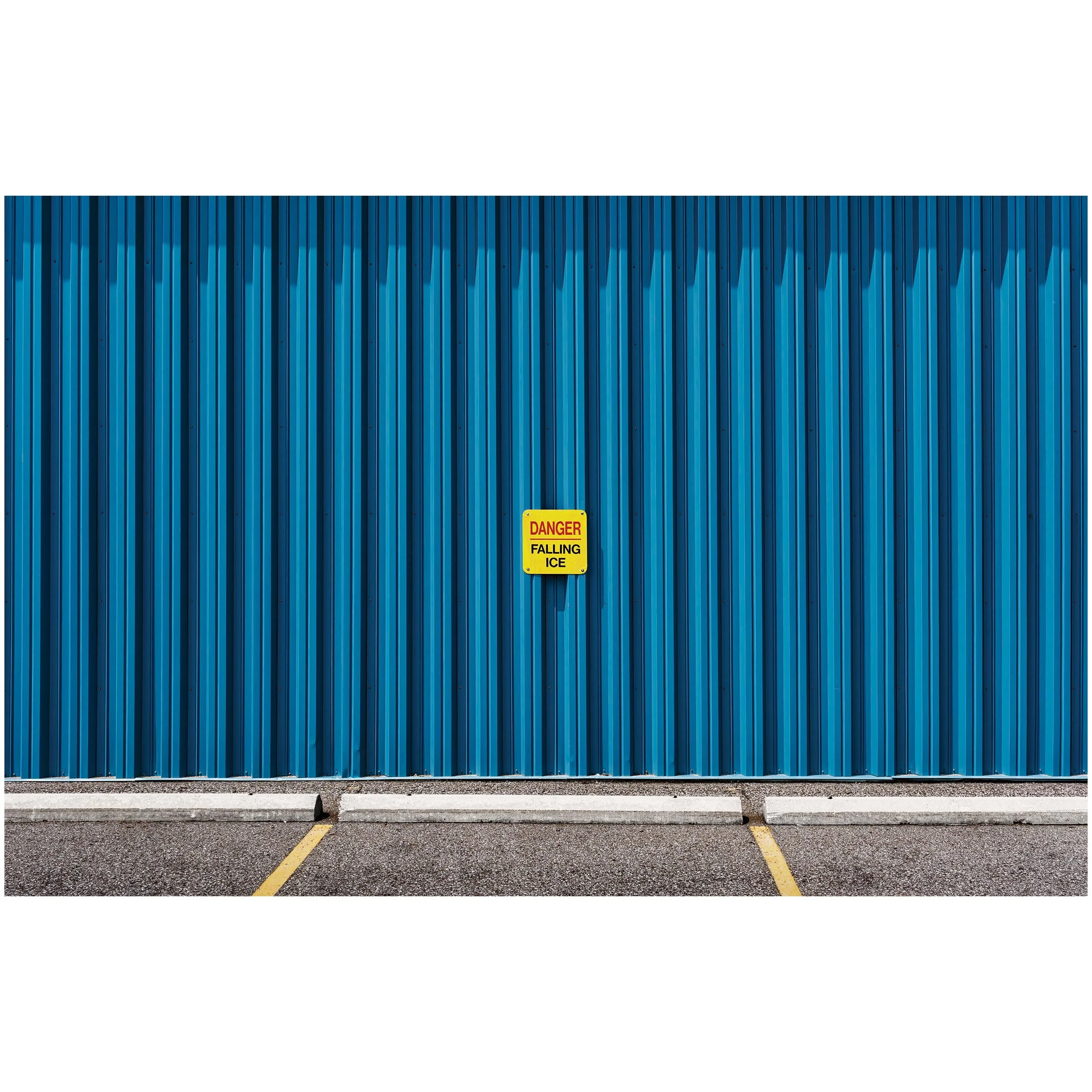 Blue metal wall with a yellow warning sign that reads "Danger Falling Ice", situated in a parking lot with yellow lines and a concrete curb.