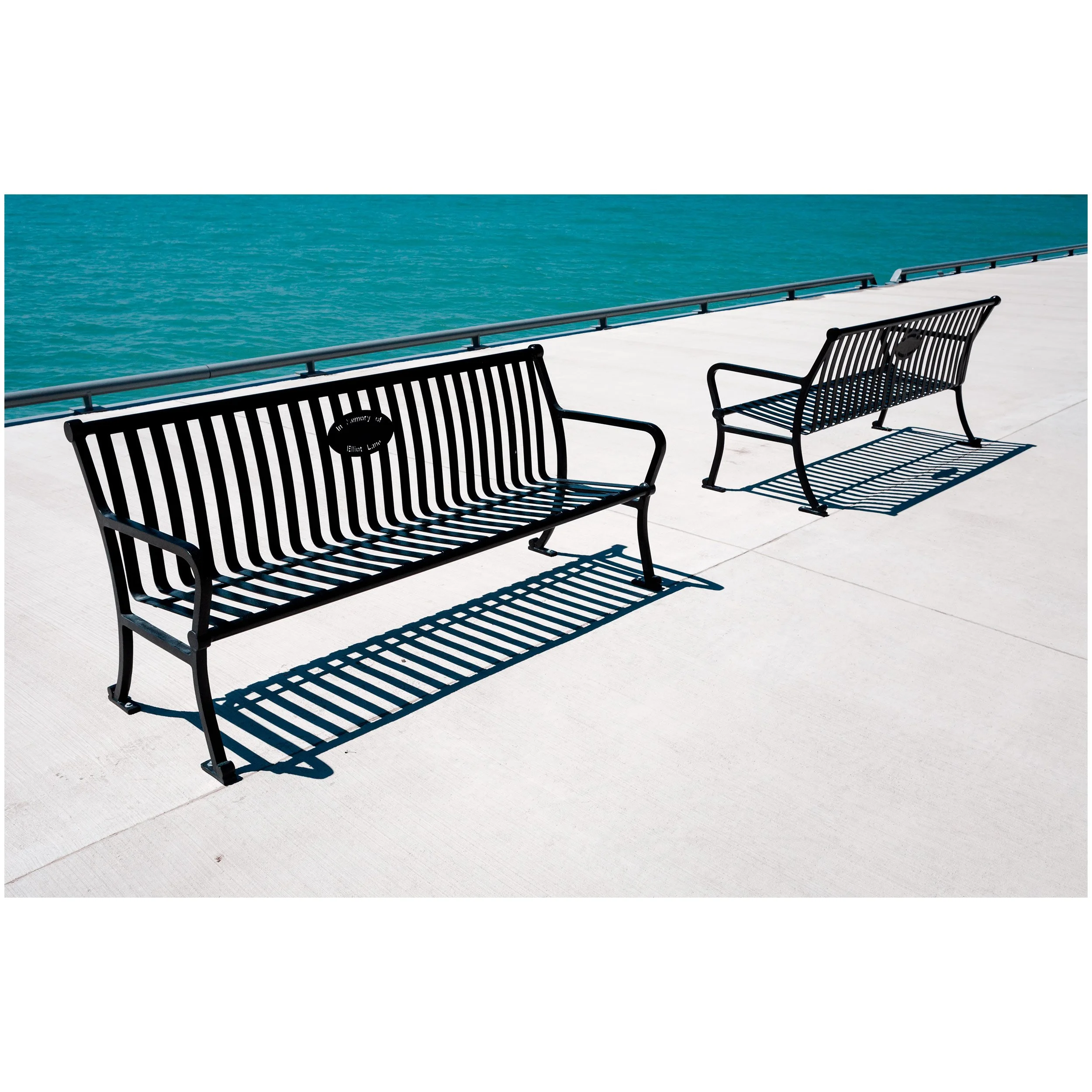 Empty black metal bench on a white concrete pier overlooking turquoise water with a clear sky.
