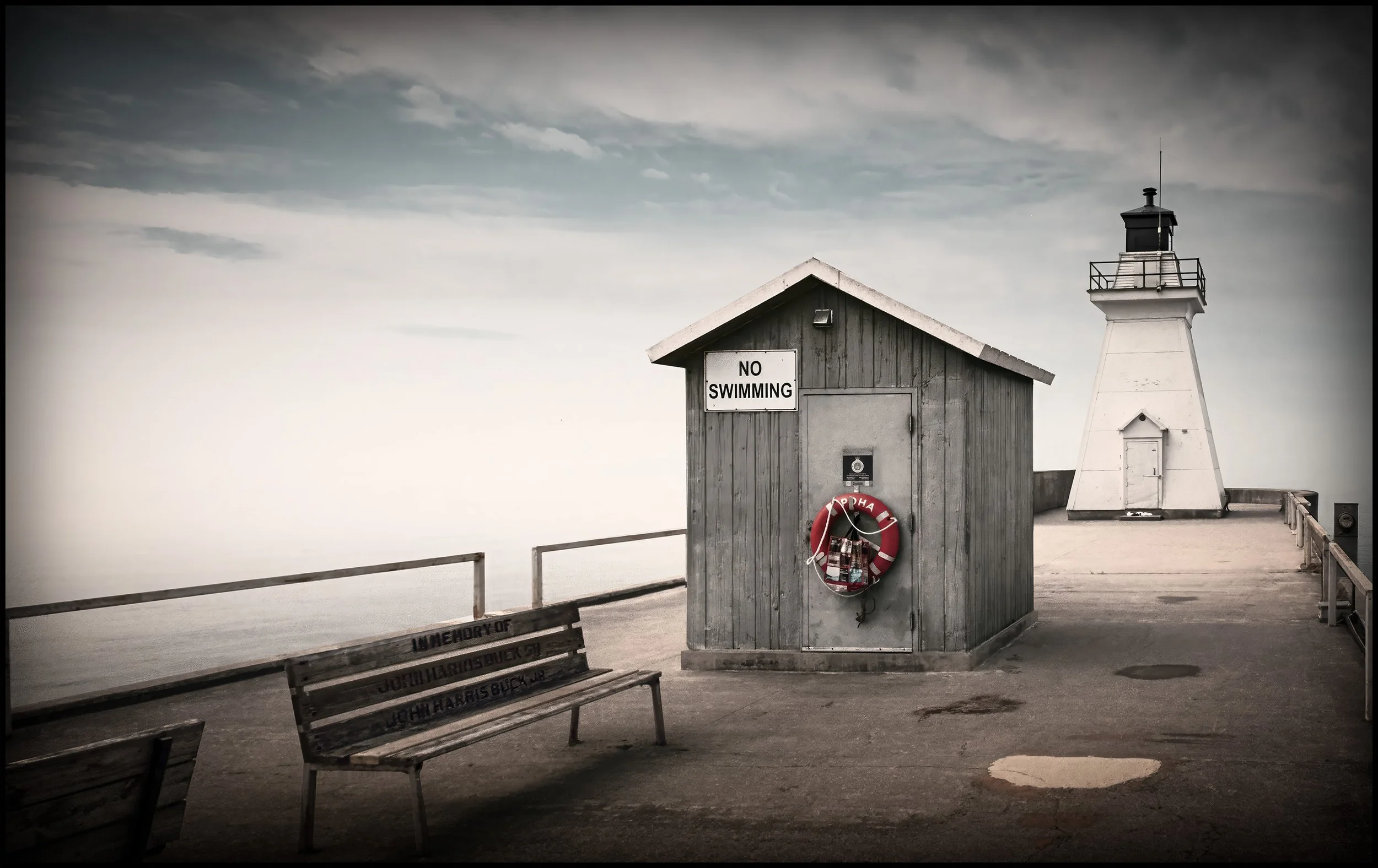 A black-and-white photo of a lighthouse and a small wooden building with a sign that says 'NO SWIMMING,' on a pier or walkway with benches and street lamps, and cloudy sky in the background.