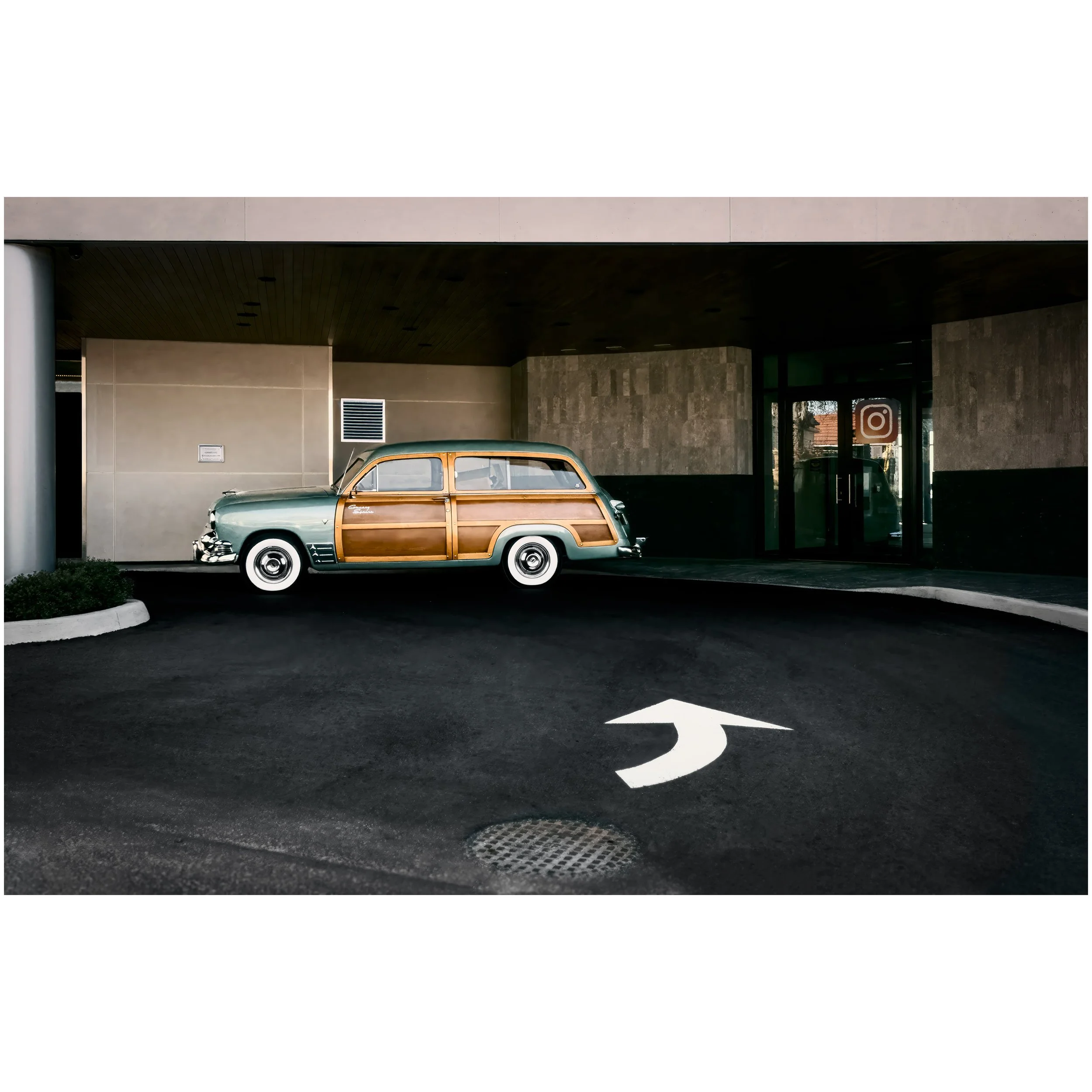 A vintage green and brown station wagon parked under a building with modern architecture, black ceiling, beige walls, and a window with an Instagram logo sticker.