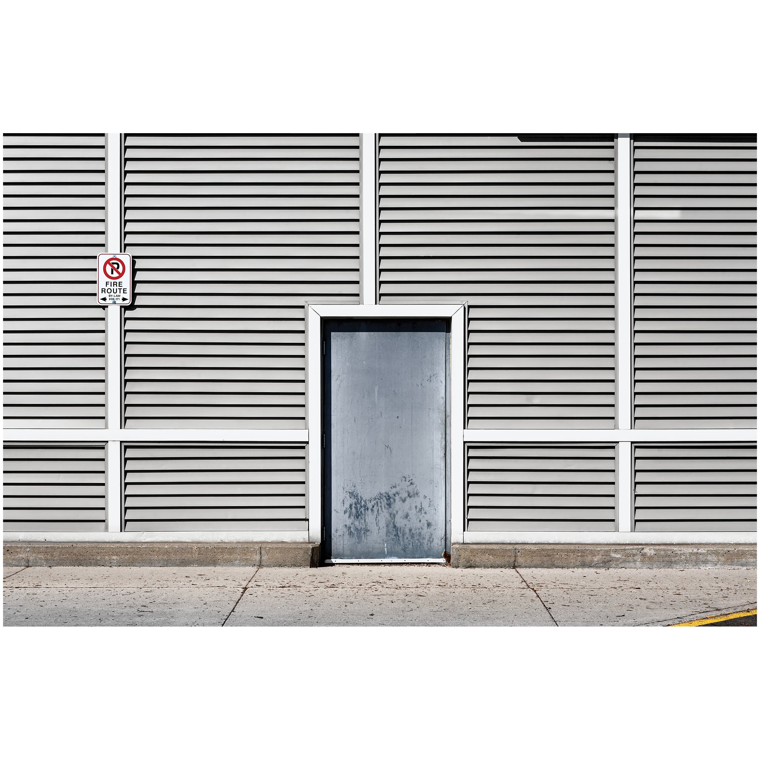 Gray building with horizontal slats and a metal door, adjacent to a sidewalk with yellow parking stripes and a no parking sign.