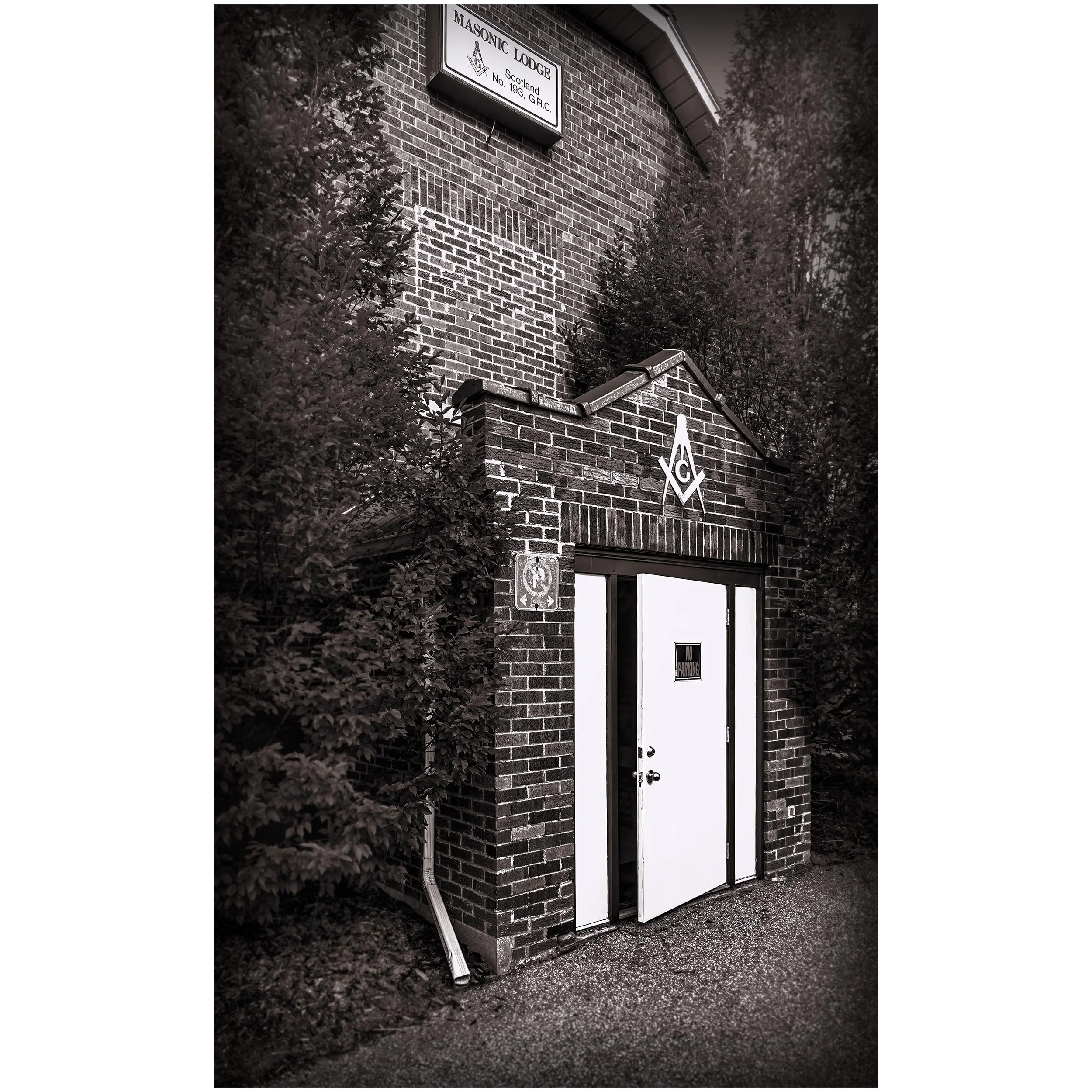 Black and white photo of the exterior of a brick building with a sign that reads "Masonic Lodge" and another sign below indicating parking restrictions, with some bushes and trees around.