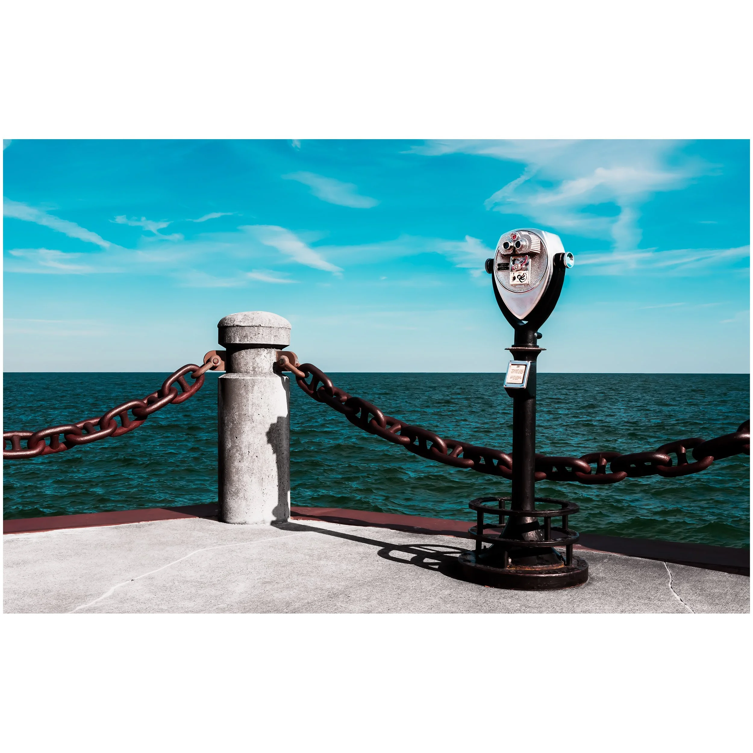 View of the ocean with a clear blue sky and some clouds. A coin-operated binocular viewer is mounted on a pole, and a chain barrier is attached to a post along the promenade.