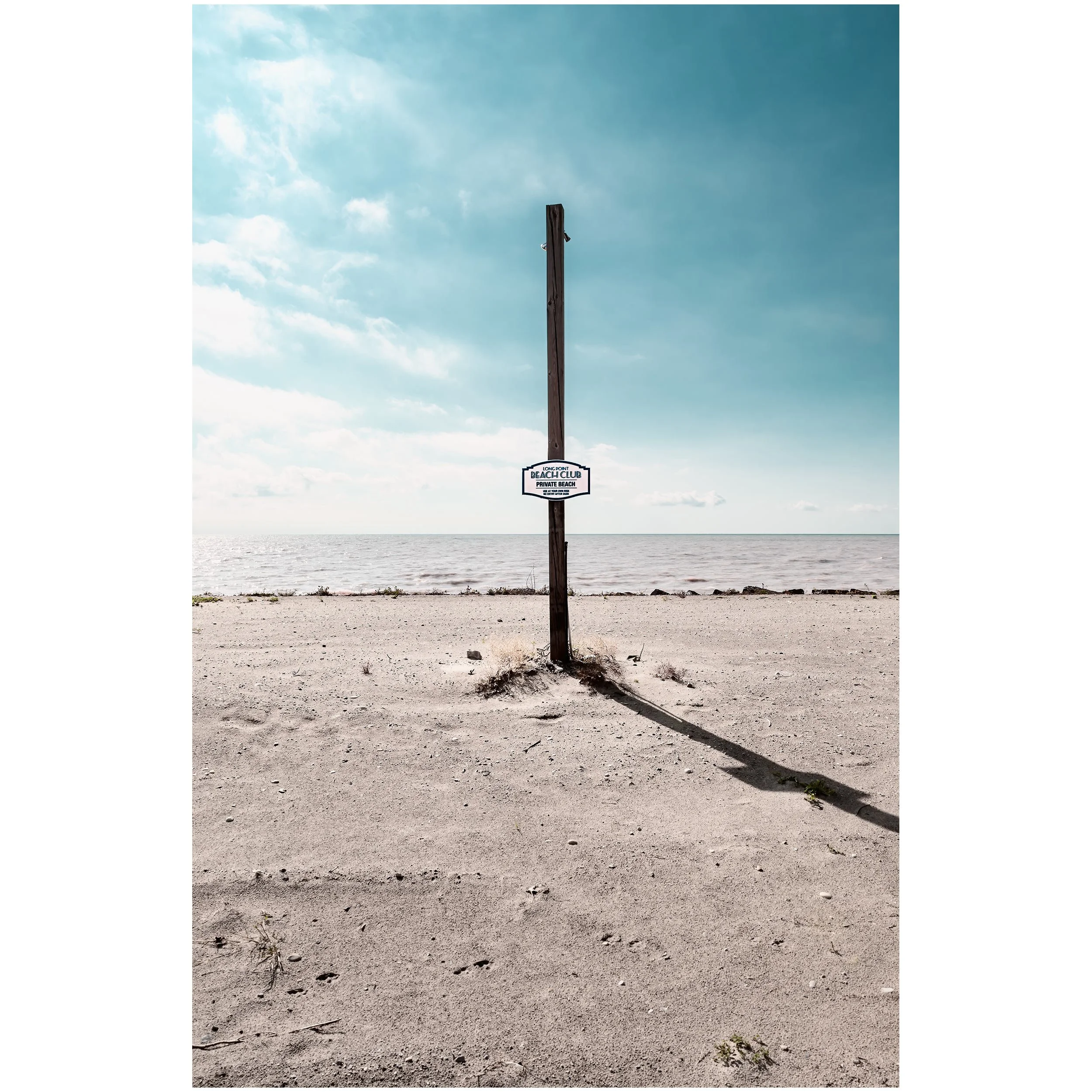 A sandy beach with some small plants, a wooden pole with a sign reading 'Long Point Beach Club Private Beach,' and a body of water in the background under a partly cloudy sky.