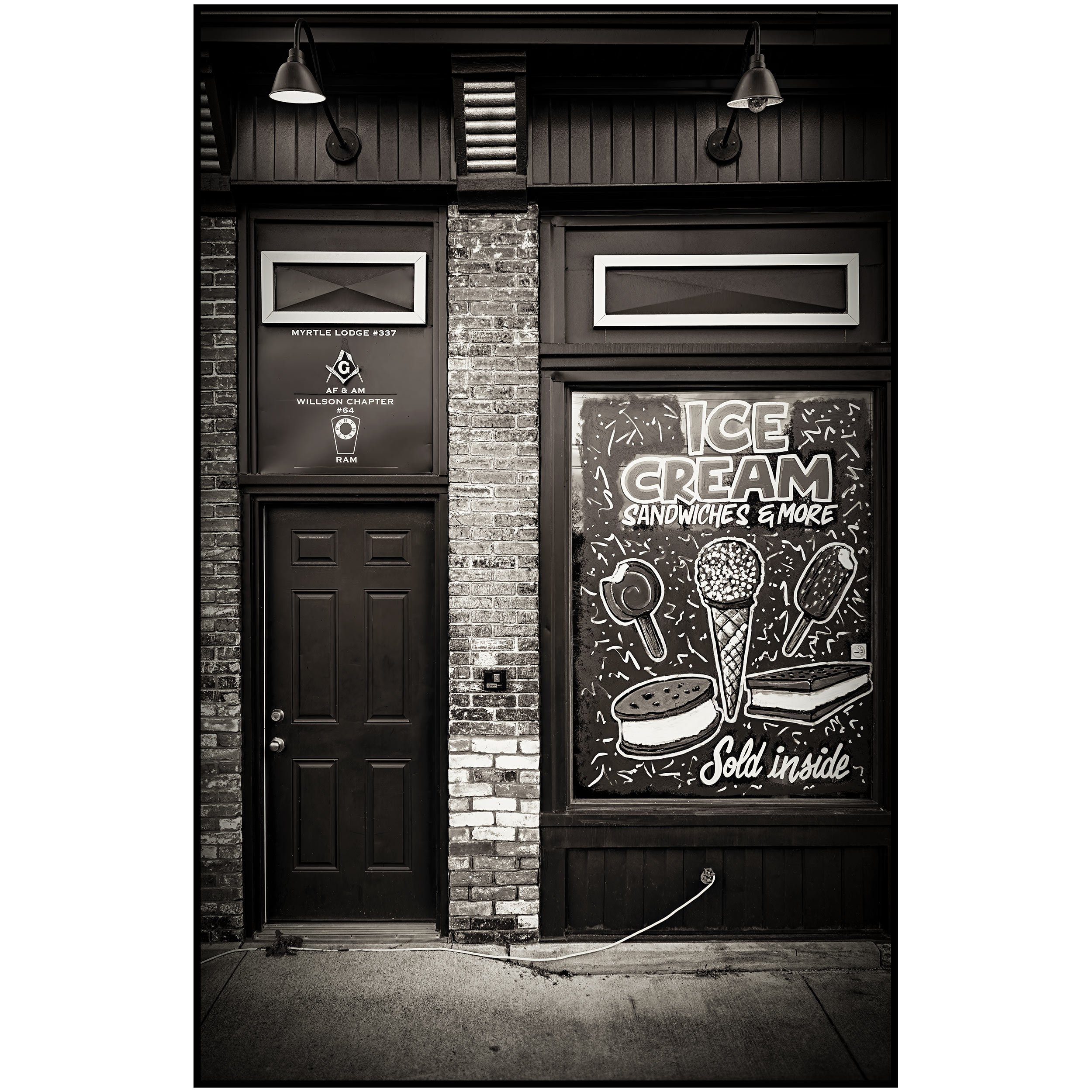 A storefront with a brick wall and black framing, featuring a mural advertising ice cream and sandwiches. The mural includes illustrations of ice cream cones and popsicles, with the text "ICE CREAM SANDWICHES & MORE" and an additional phrase "Sold inside." Two black wall-mounted lamps are positioned above the mural, illuminating the storefront.