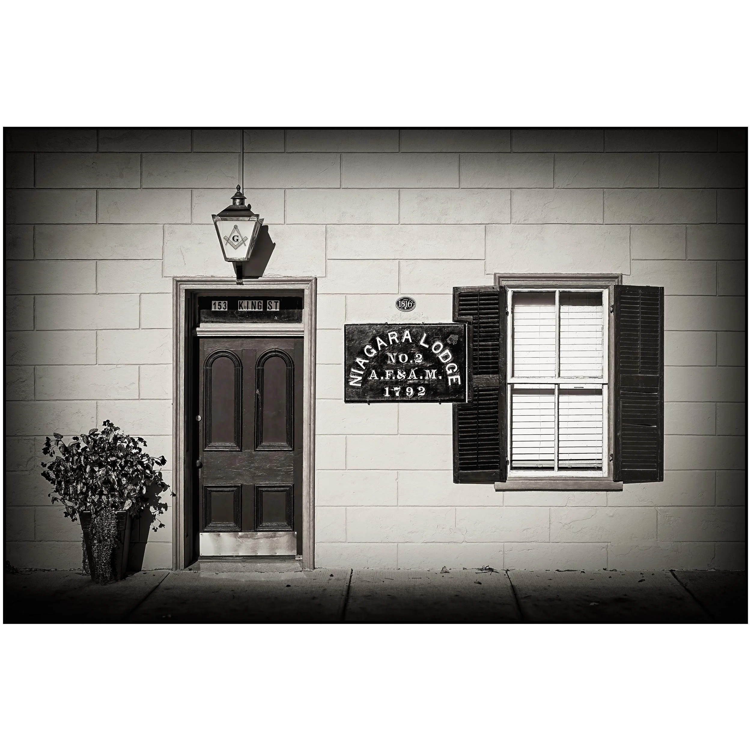 A black and white photo of a white brick building with a front door, a window with shutters, a hanging lantern with a Freemason symbol, and a sign that reads 'NINETEEN CAR LODGE NO. 2 A.F.&A.M. 1792'. There is a potted plant in front of the door.
