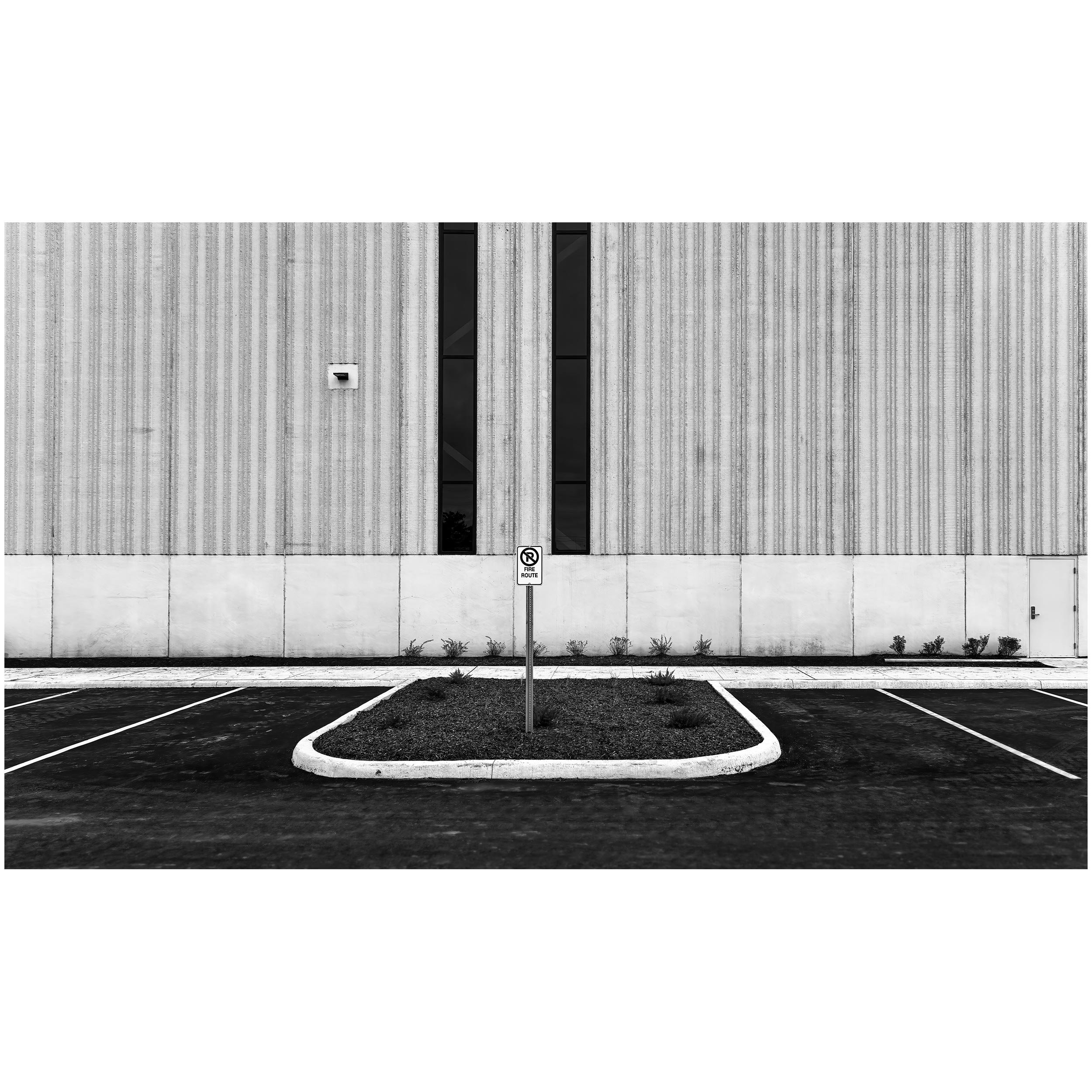A modern building facade with vertical striped concrete or stone panels, two narrow tall windows, and a parking lot with marked spaces in front, including a no-parking sign for fire routes.