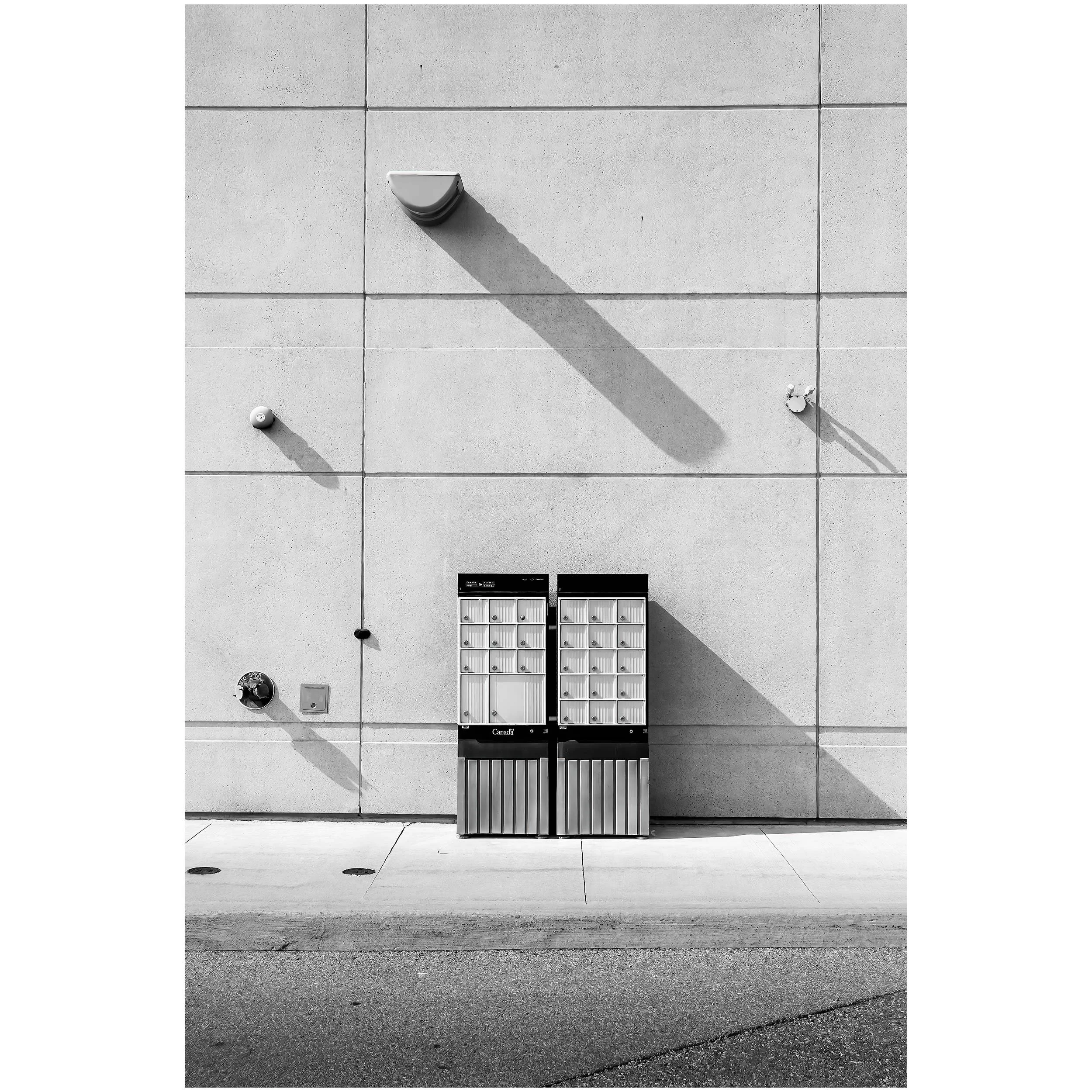 Black and white photo of a building's exterior wall with utility fixtures, including a vent, a security camera, and an air conditioning unit, casting shadows on the wall.