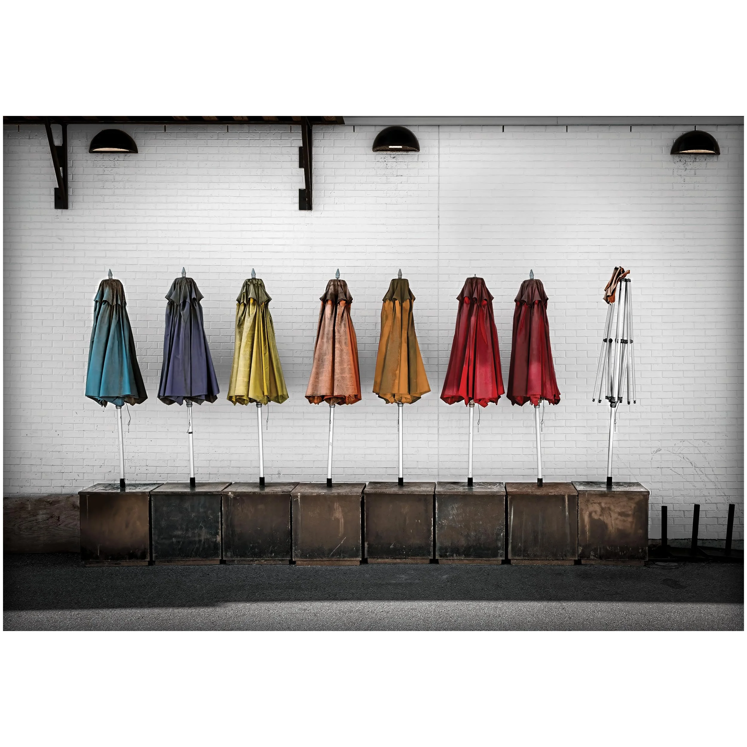 Display of old colourful closed umbrellas against a white brick wall, with a black platform and a ladder on the right side.