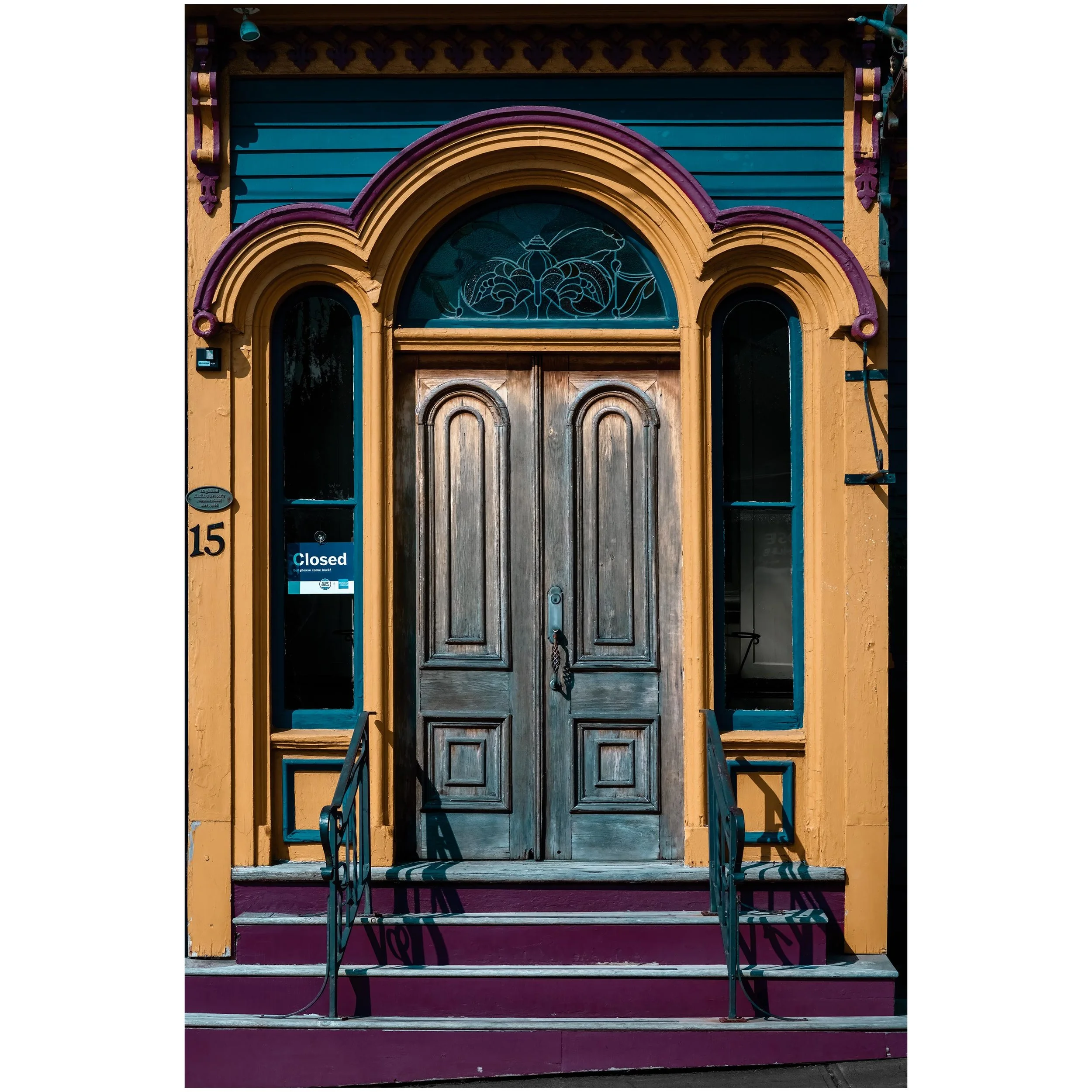 Colorful building facade with a wooden door beneath a decorative arched window, painted in yellow, teal, purple, and pink. Metal stairs lead up to the door, and a small sign indicates the business is closed.