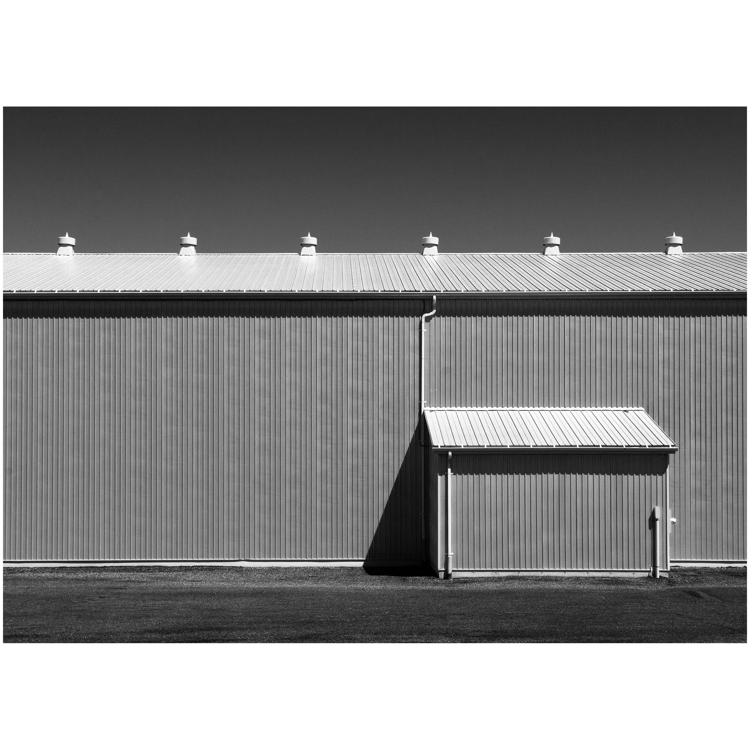 Black and white photo of a building's metal roof with multiple vents and a pipe, set against a dark sky and textured ground.