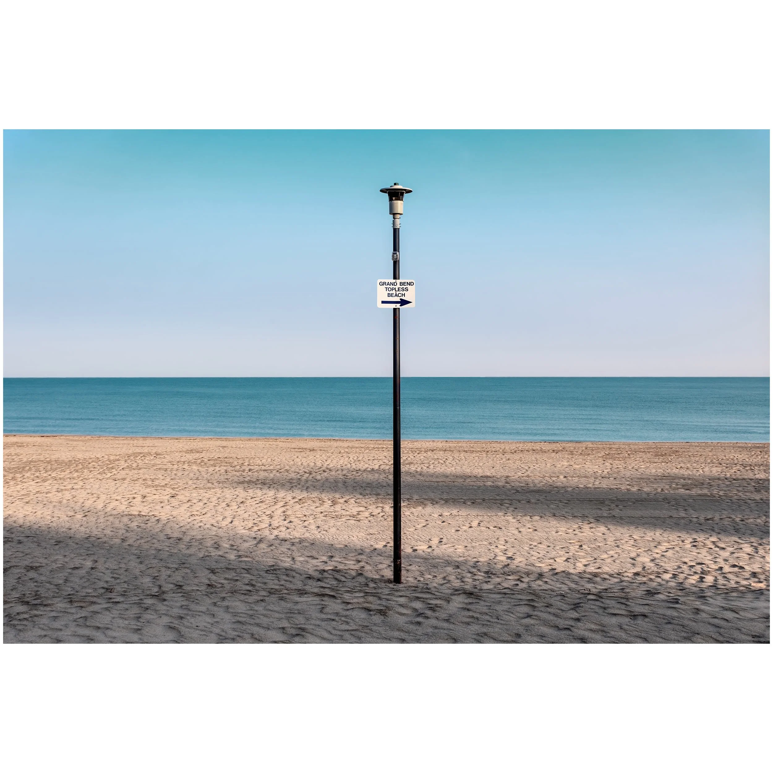 A beach scene with a sign that reads 'Grand Bend Topless Beach' and an arrow pointing to the right, set against a clear blue sky, calm ocean, and sandy shore.