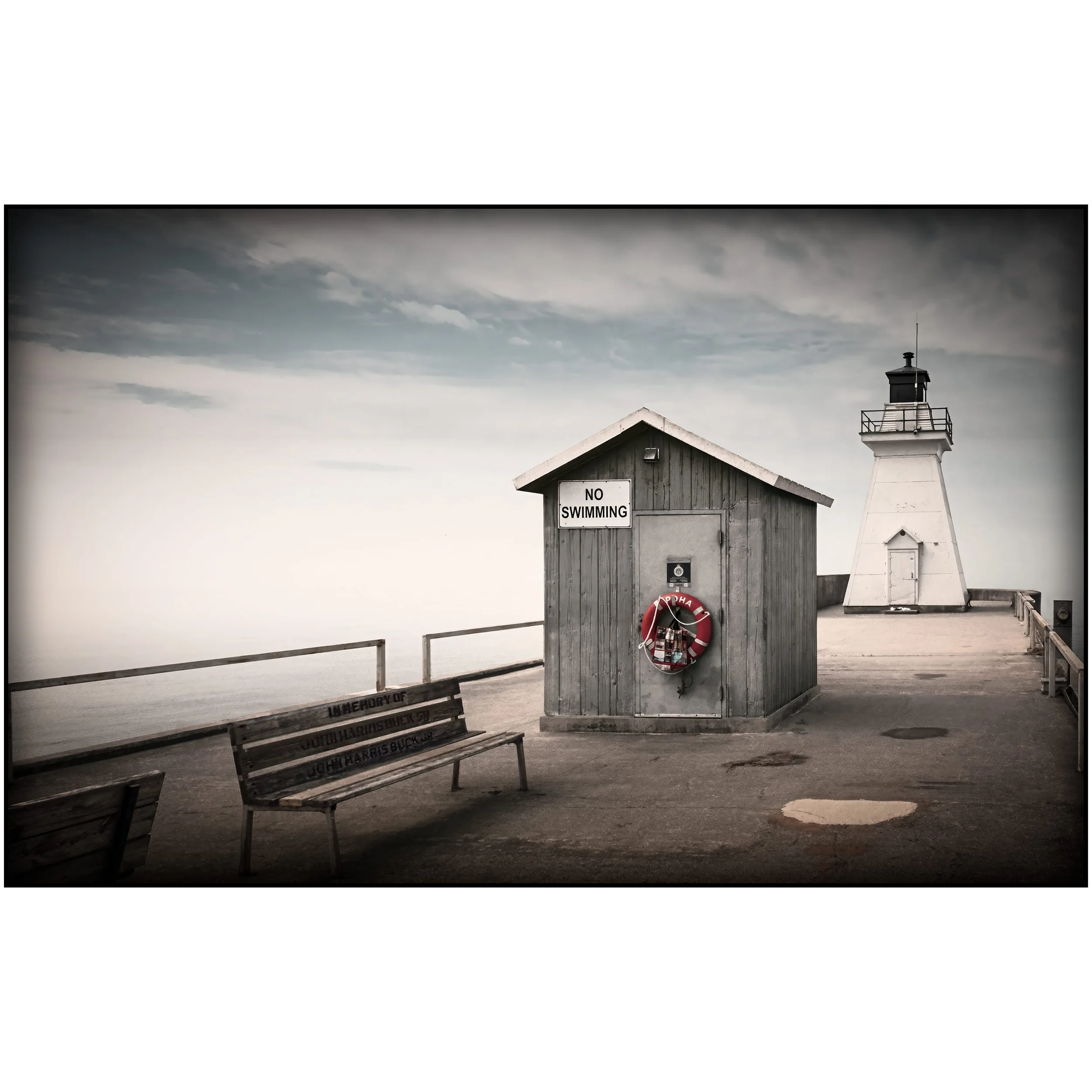 A weathered wooden shed marked “No Swimming” stands on a quiet pier beside benches and railings, with a white lighthouse behind it under a muted sky.