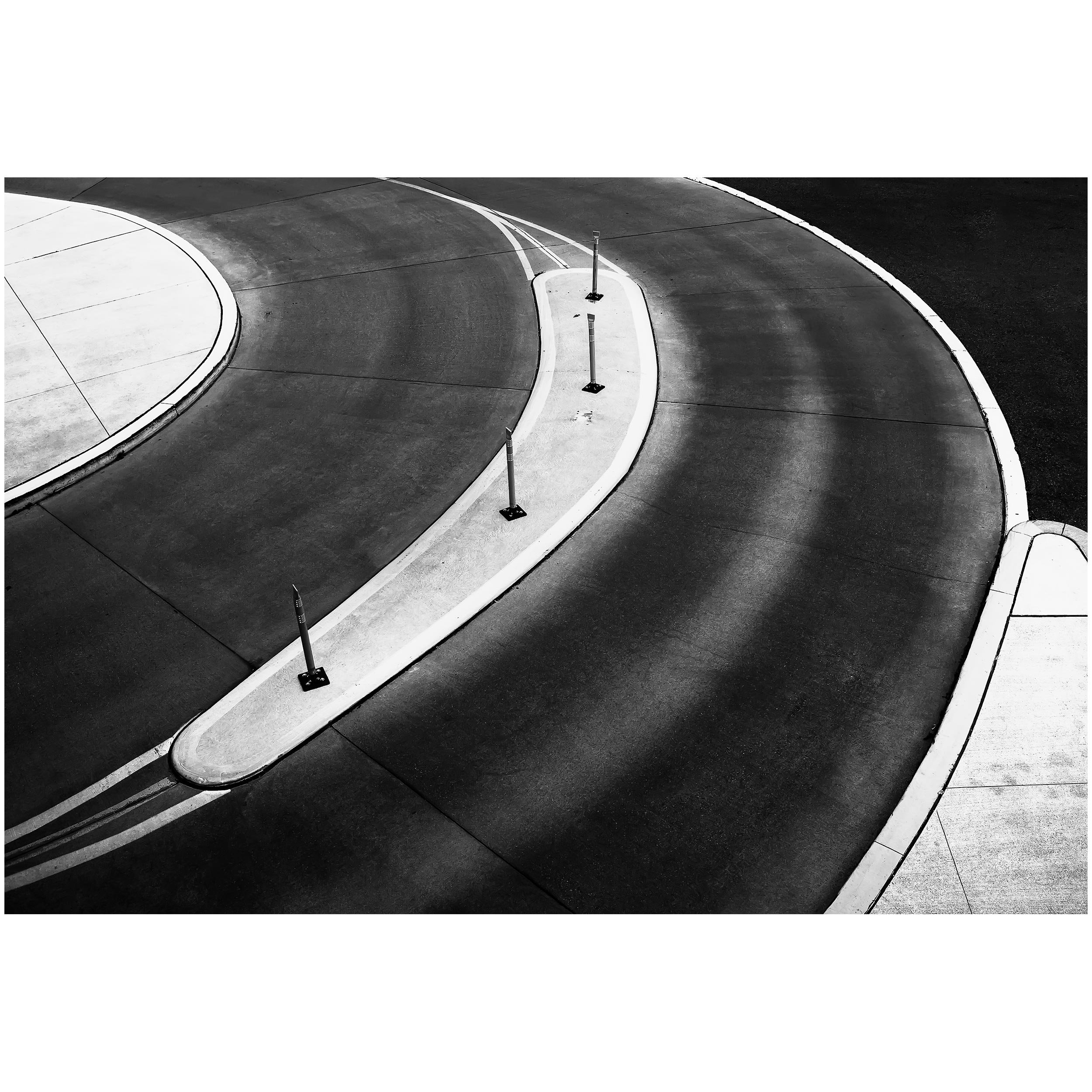 Black and white image of a curved parking lot with small poles along the edge and painted lines on the asphalt.