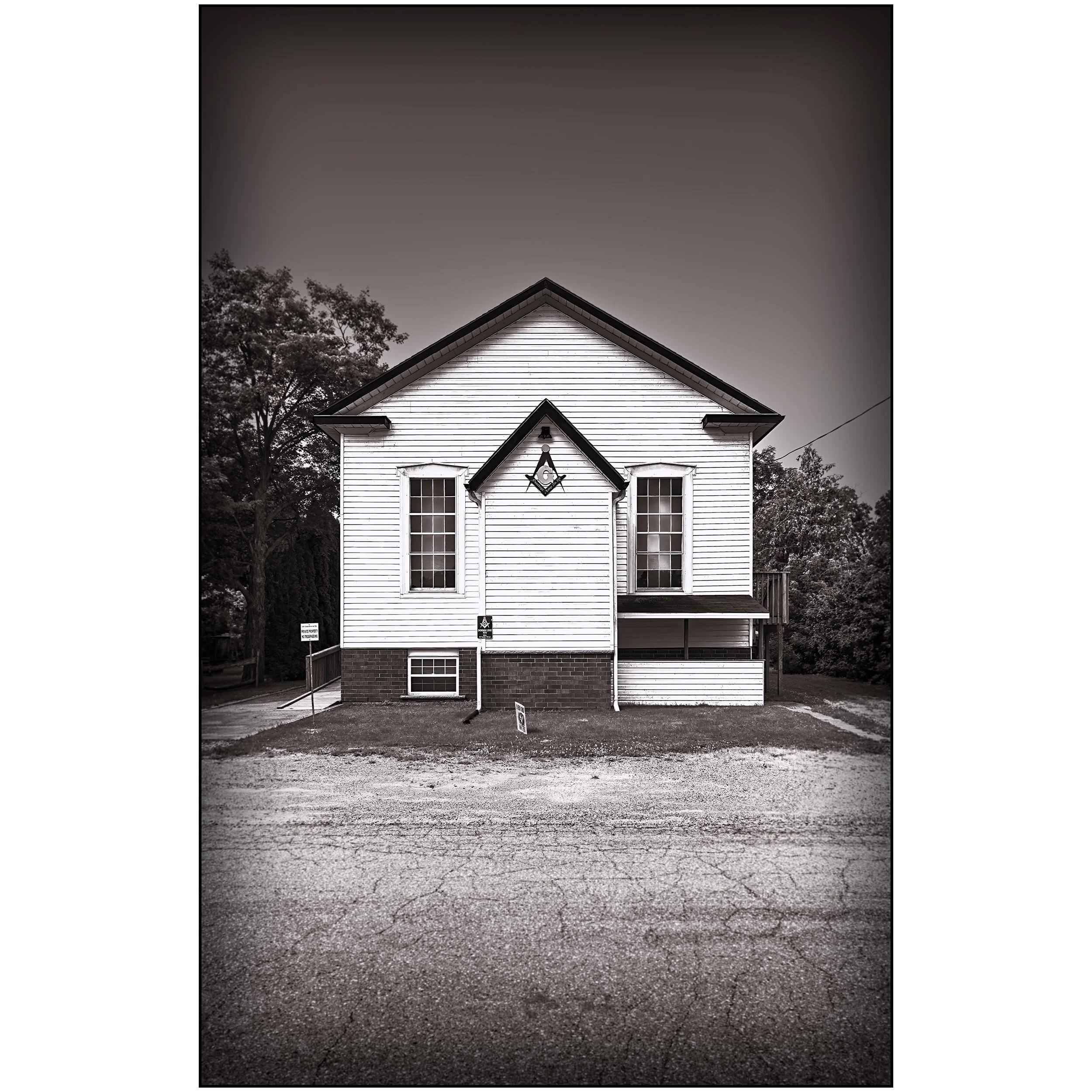 A black and white photo of the front of a white wooden house with two tall windows, decorative trim, and a truss design on the front. There are trees on the sides, and a gravel driveway in front, with some signs placed on the ground.