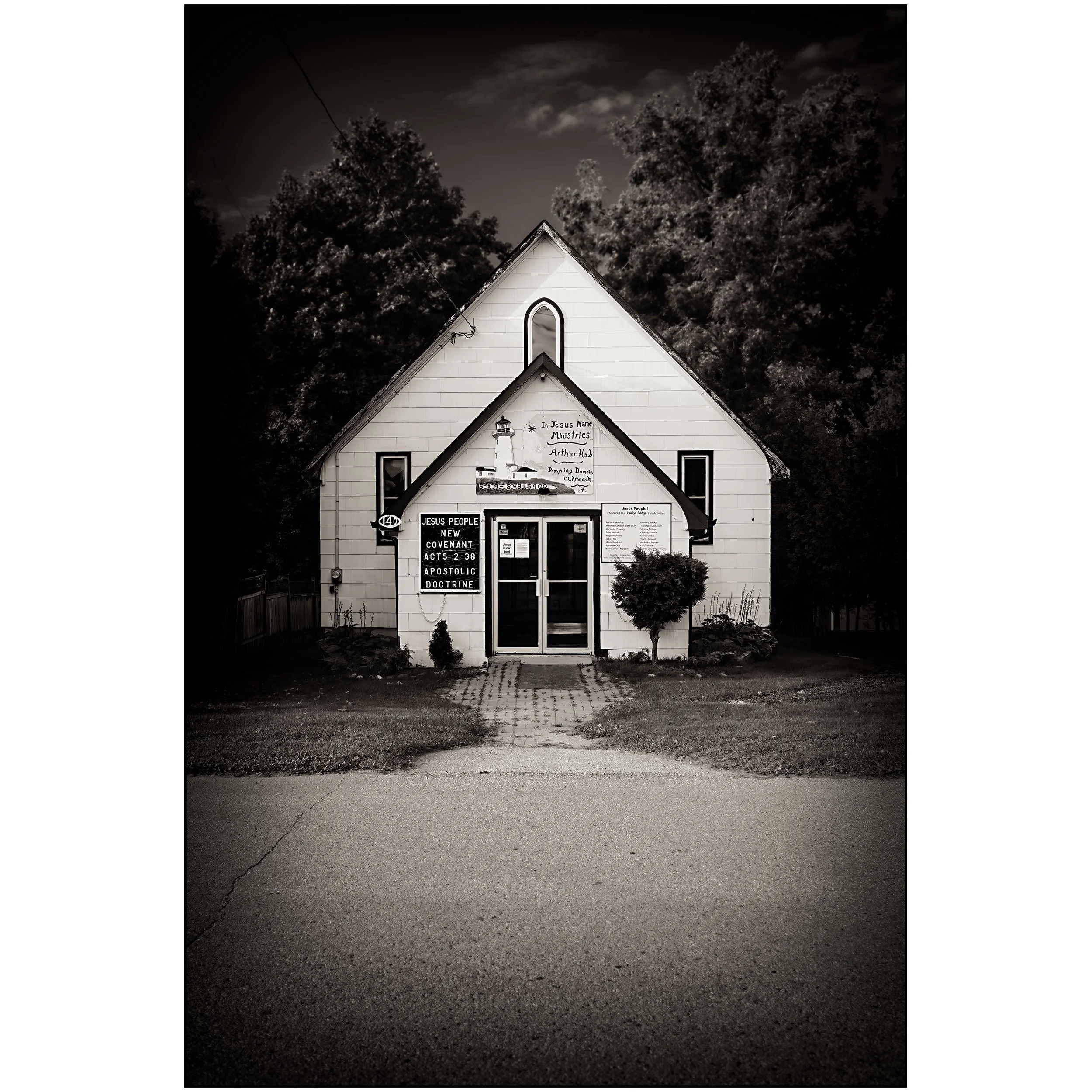 A small church with a sign that reads 'In Jesus Name Ministries Arthur Hub.' The sky is overcast and the building is surrounded by bushes. There is a parking lot in front of the church with a few cars parked.