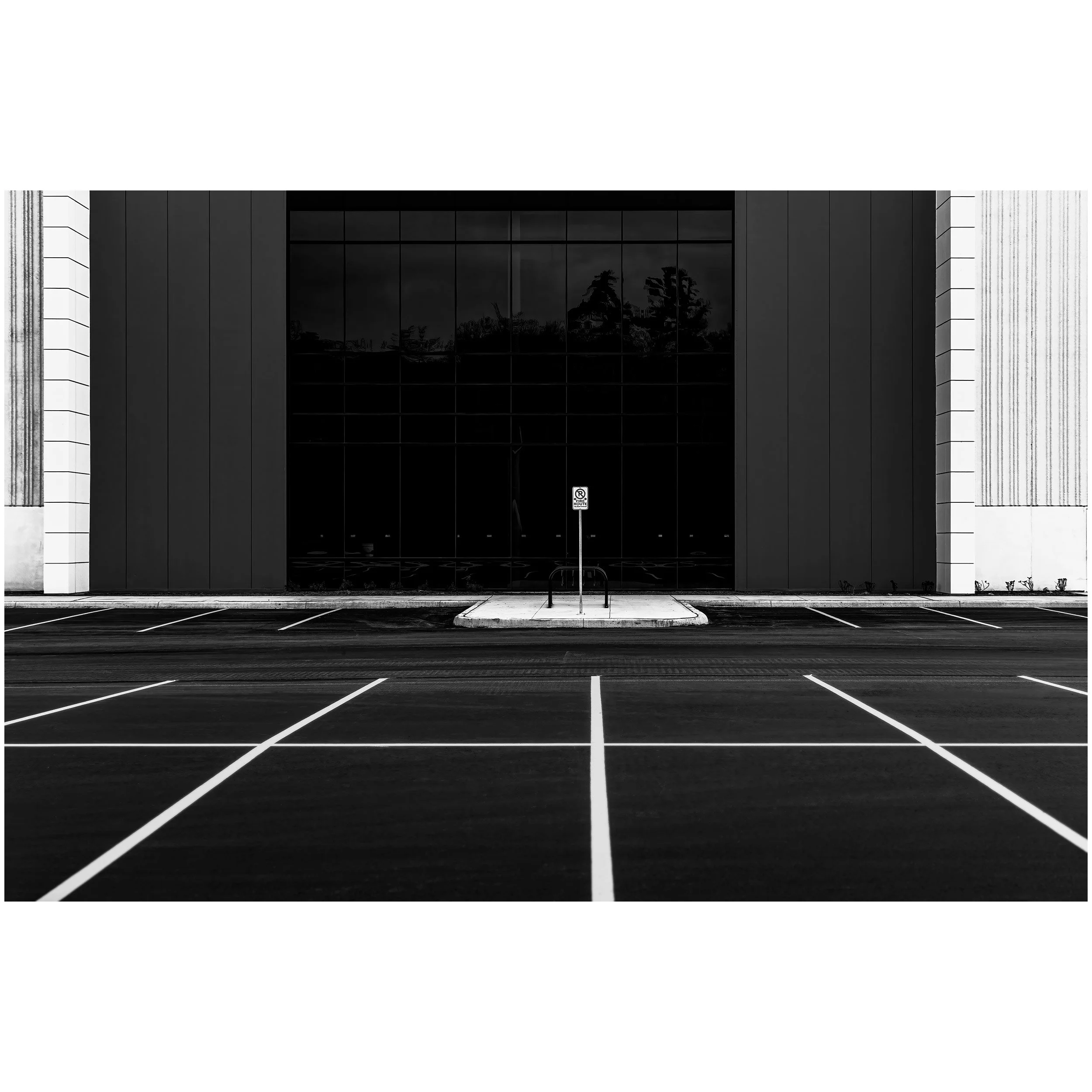 Empty parking lot in front of a modern building with reflective glass windows, black and white exterior, and trees reflected in the glass.