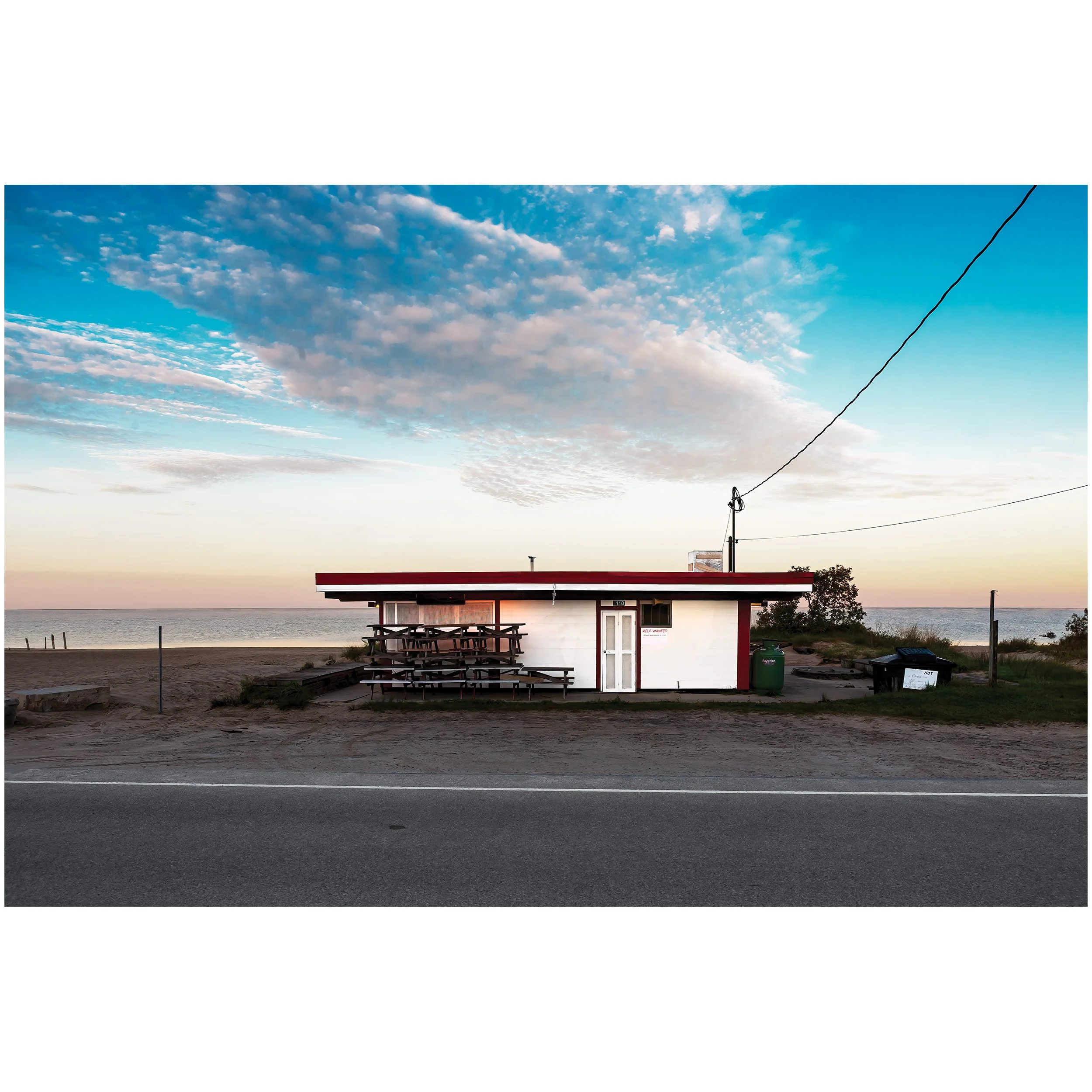 Photo of a small, white building or kiosk with a red stripe along the roof, situated near a body of water, with a clear sky overhead featuring scattered clouds. There are long picnic tables in front of the building and overhead wires crossing the scene.
