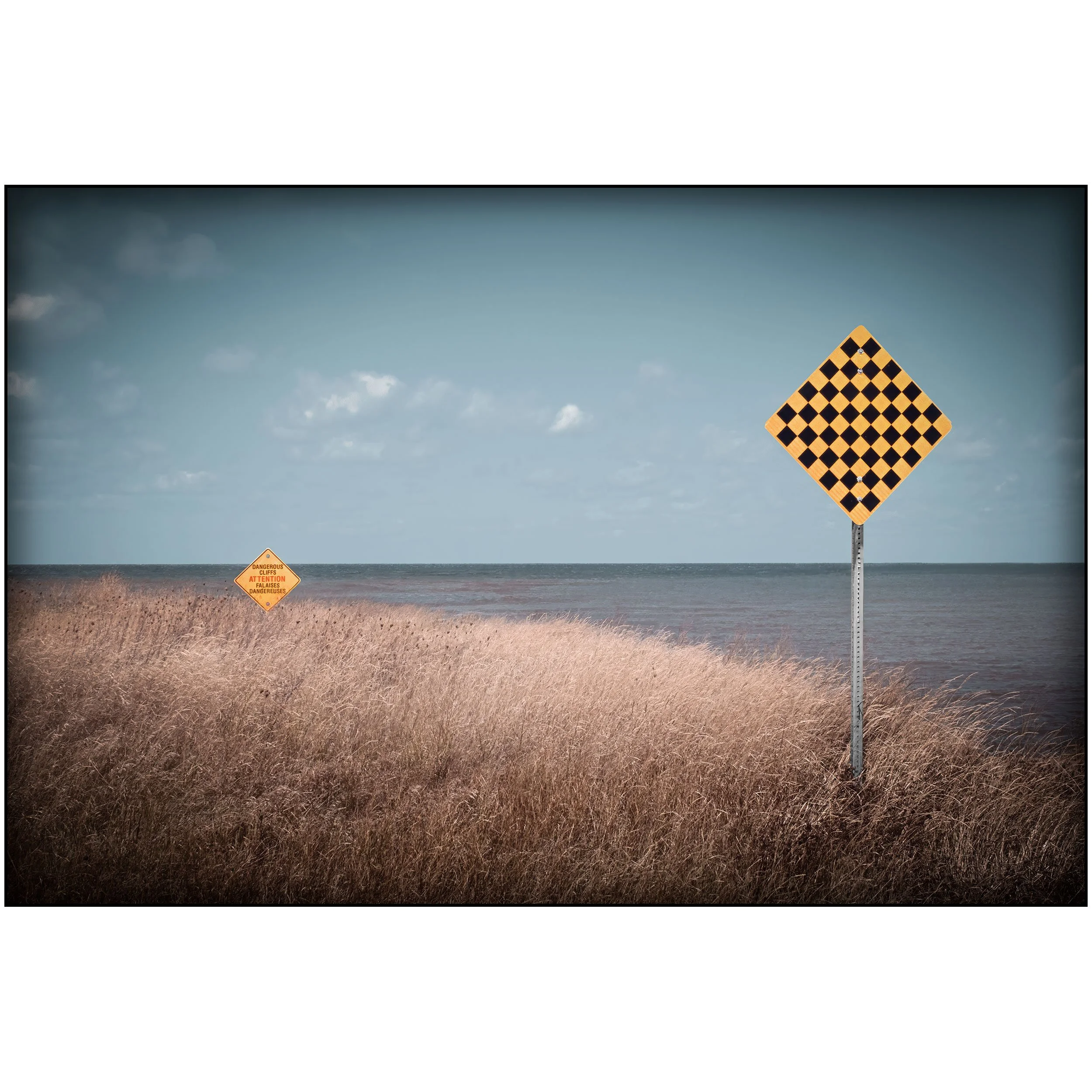 A triangular warning sign with a black and orange checkerboard pattern in a field of dry grass, and a smaller warning sign in the background reading 'DANGEROUS CLIFFS' in orange and black.