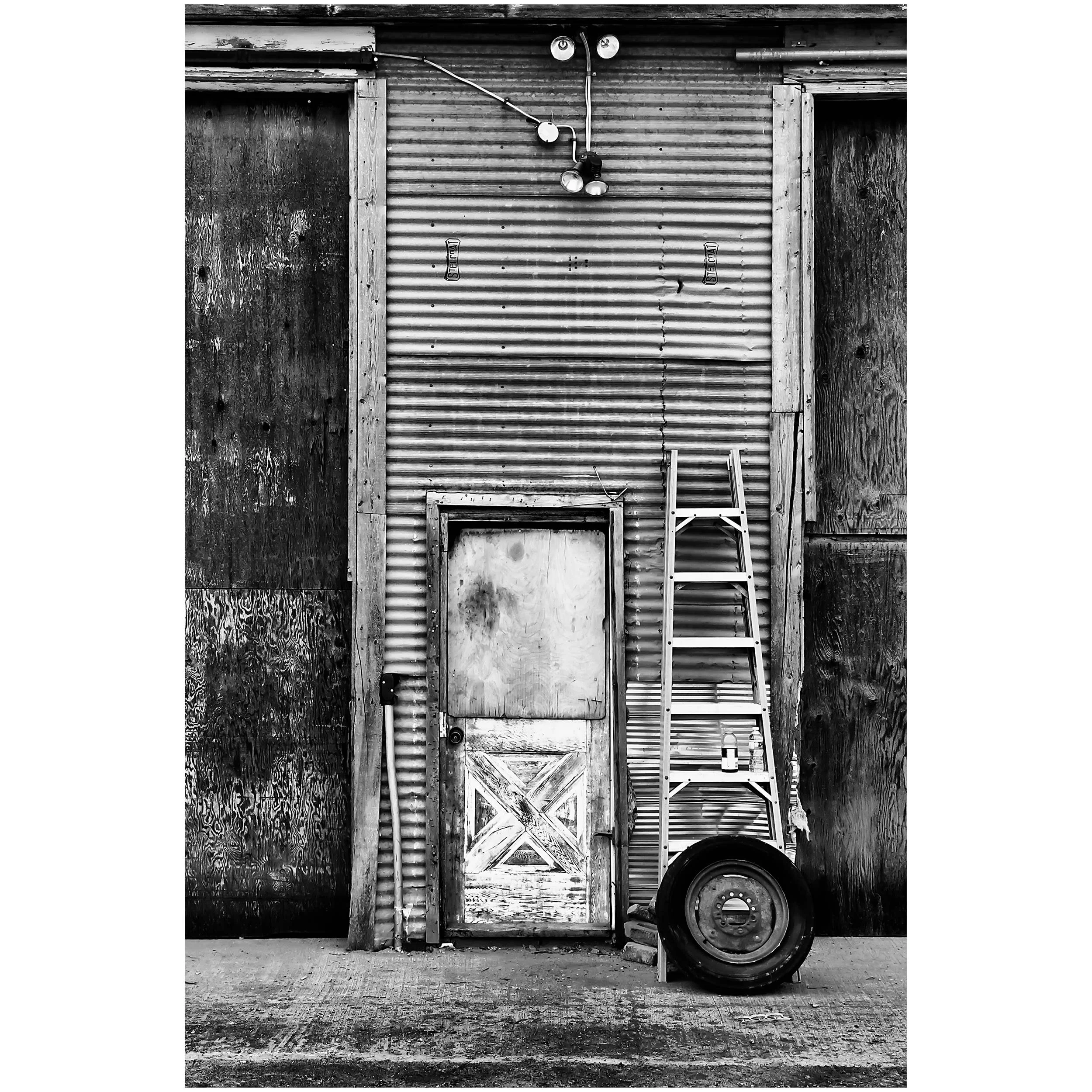 Black and white image of a rustic, weathered building exterior with corrugated metal siding, wooden framing, a ladder, a tire, and utility lights.