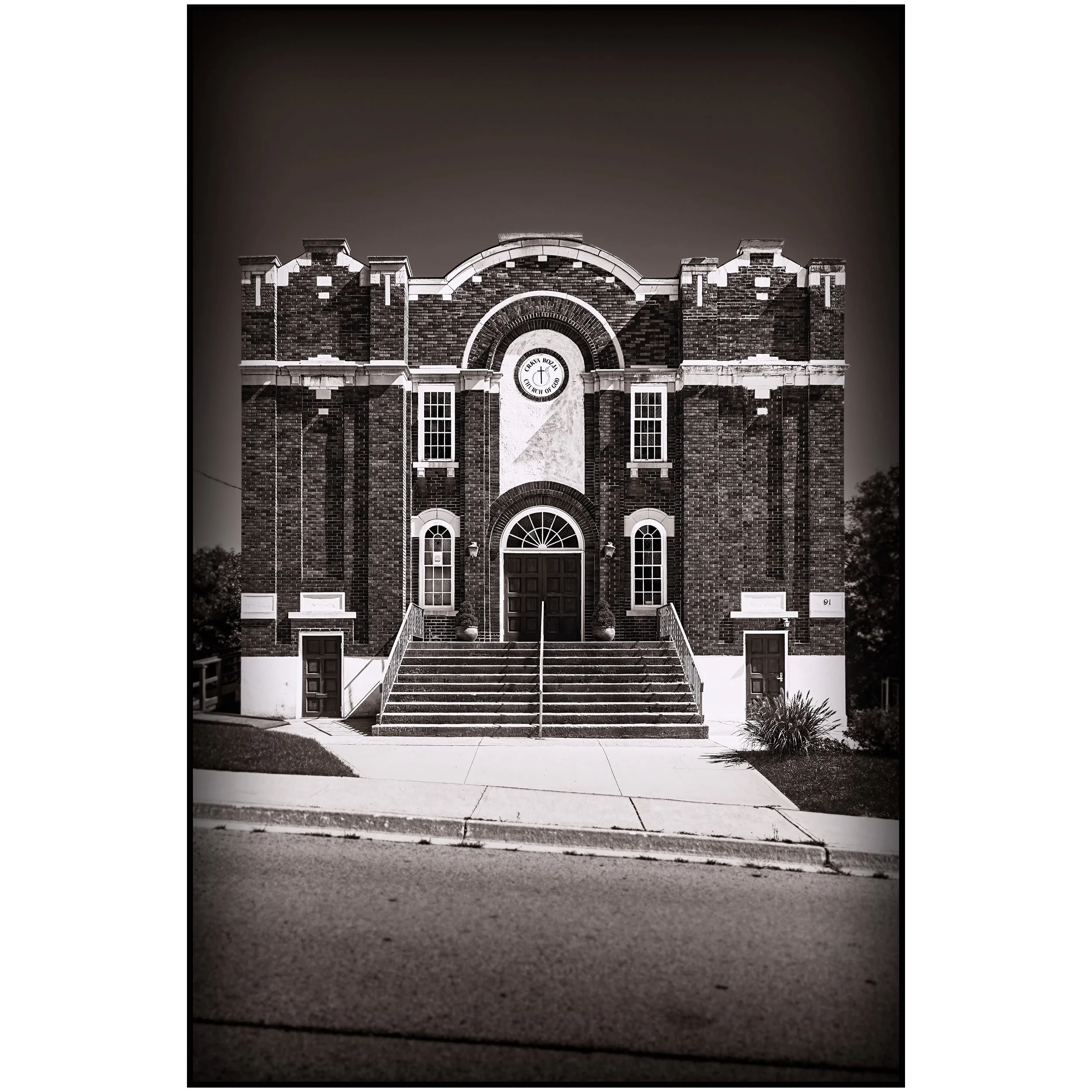 Black and white photo of a historic brick church with arched windows and a circular sign above the entrance that reads 'Church of God.' The church has a set of stairs leading up to the door, flanked by potted plants, and is surrounded by manicured lawns and shrubbery.