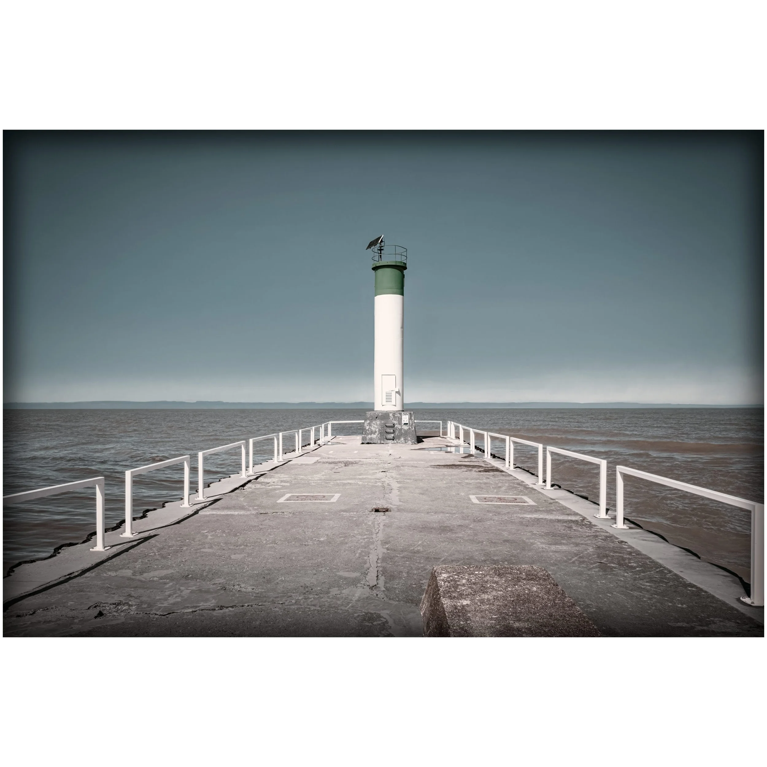 A white and green lighthouse stands at the end of a long concrete pier, framed by white railings and open water on both sides.