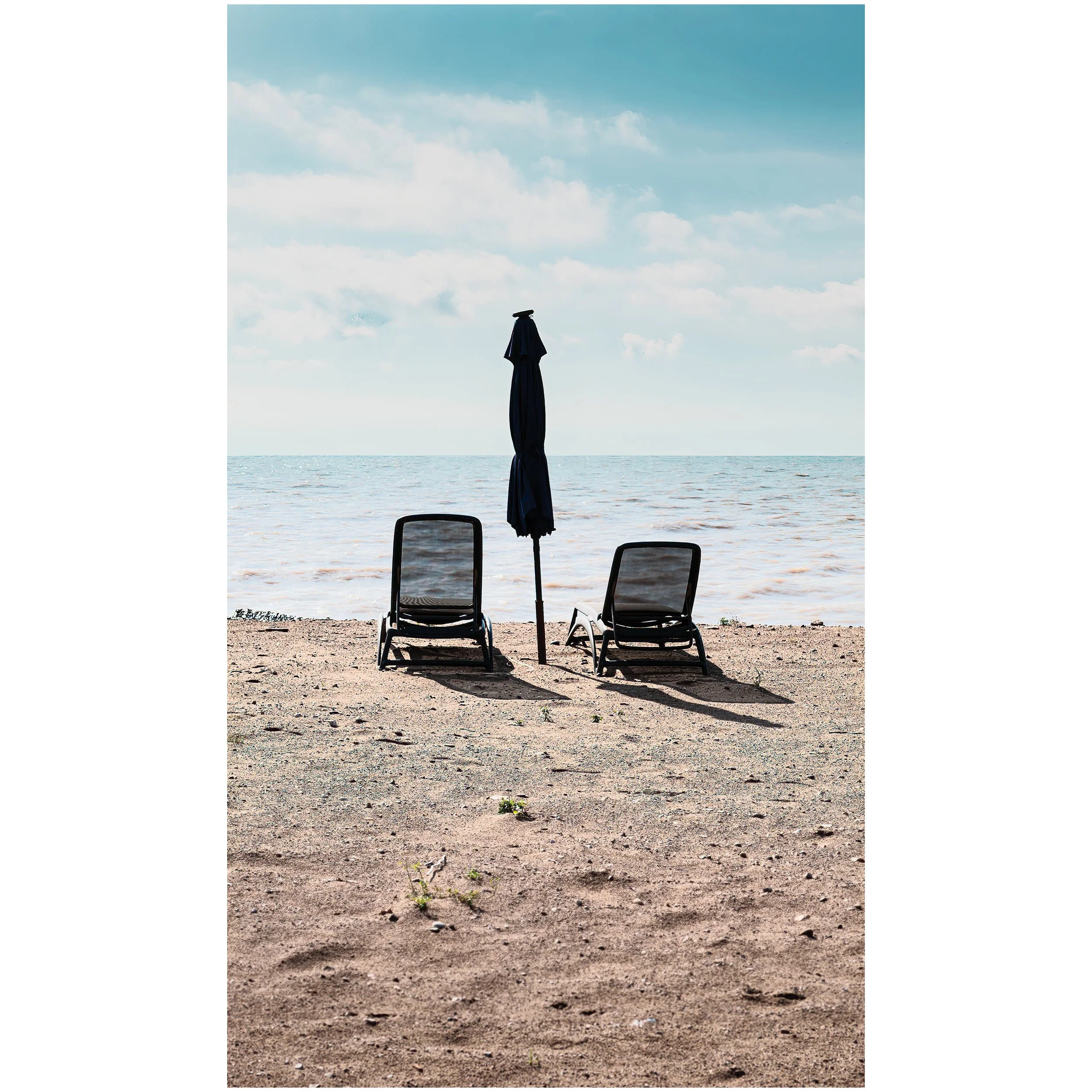 Beach scene with two empty chairs and a closed umbrella facing the ocean, with a partly cloudy sky.