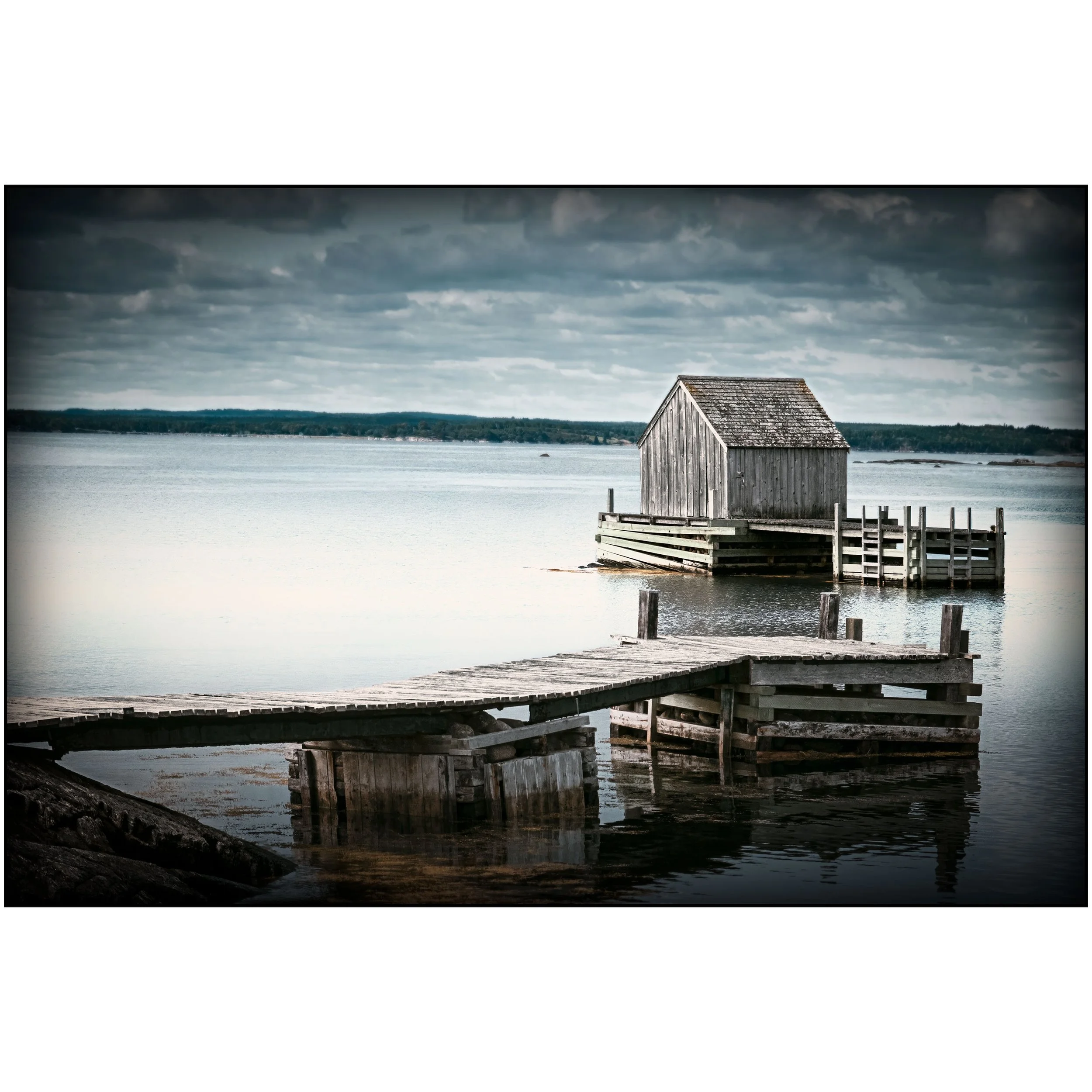 An old wooden dock stretches over still water toward a grey boathouse on pilings, with a dark cloudy sky and distant shoreline behind it.