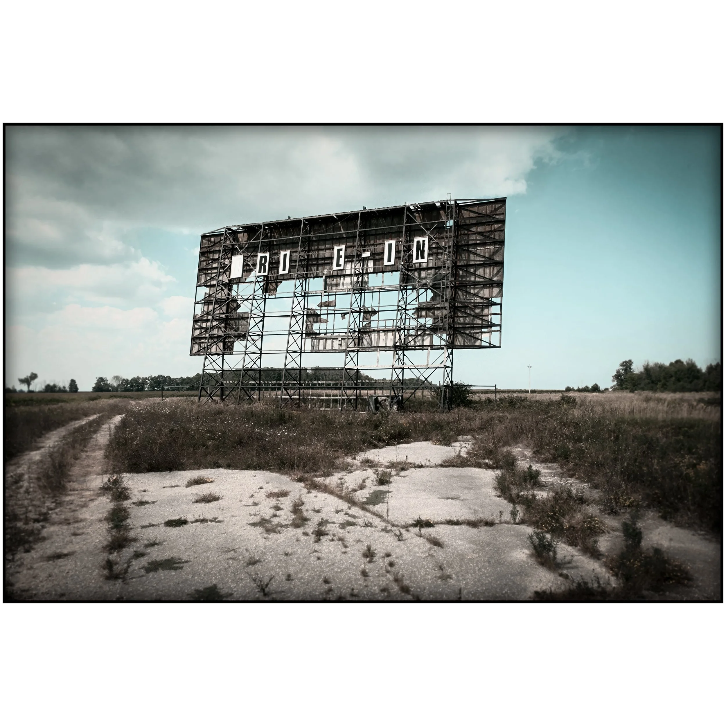 A weathered drive-in movie screen rises above cracked pavement and tall weeds, with faded letters and an open field behind it.