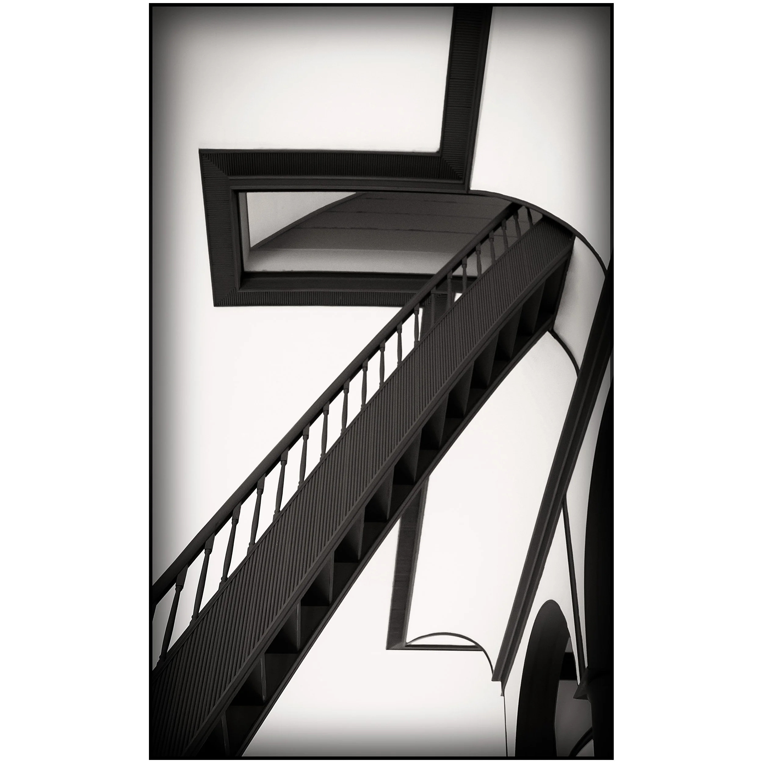 Close-up of a dark wooden staircase with railing leading upwards beneath a stairwell with a dark overhang against a plain white wall.