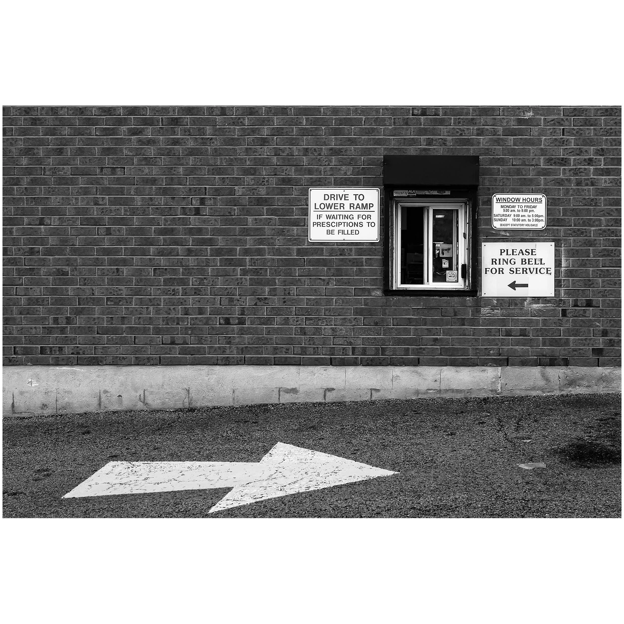 Black and white photo of a brick wall with signs, a lower ramp drive, and painted parking lines in the foreground.