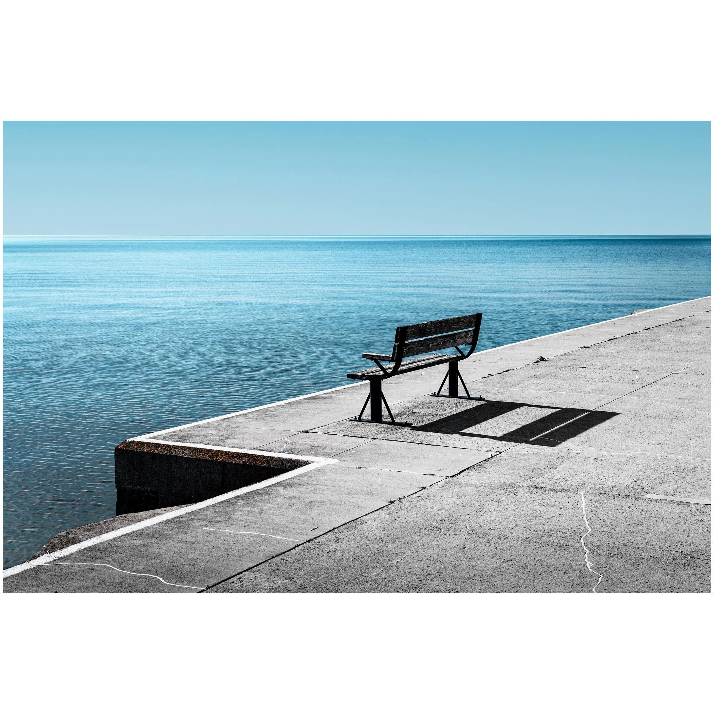 A concrete pier with benches and a railing along the water's edge, extending into a calm body of water under a clear blue sky.