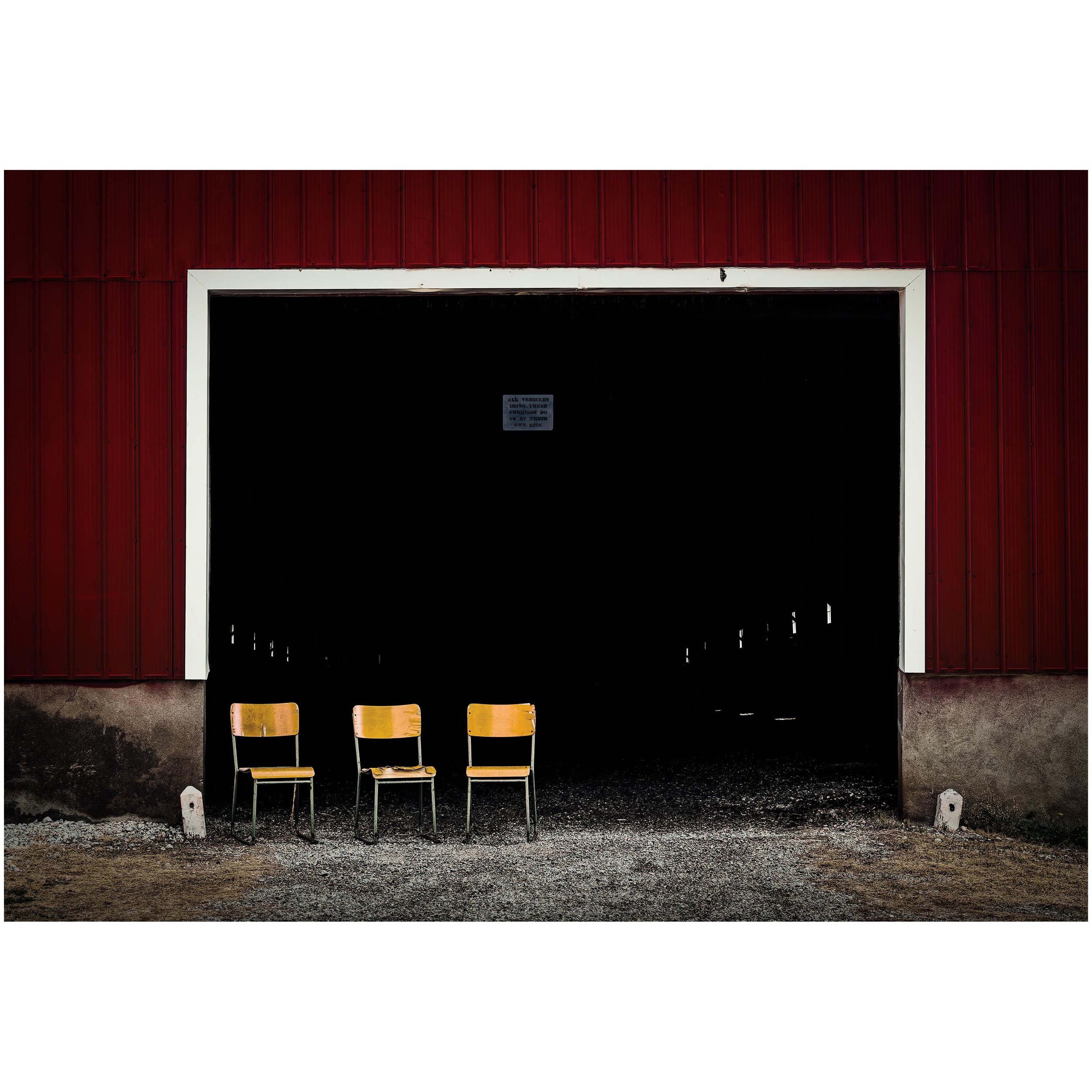 Three yellow chairs arranged in a row on a gravel ground outside a red barn with a black open doorway.