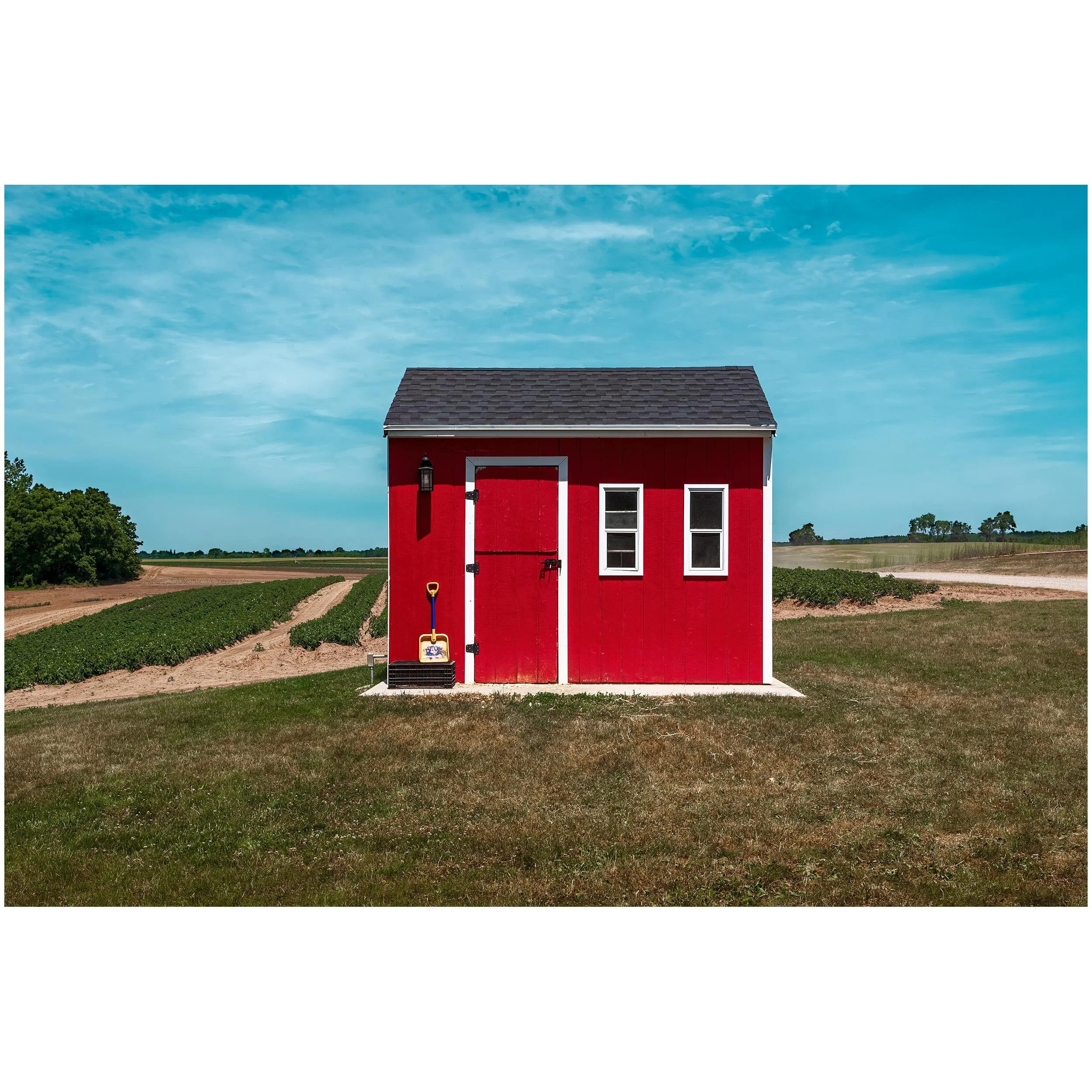 A large red barn with a black shingled roof surrounded by farmland, fields, and green trees under a partly cloudy blue sky.