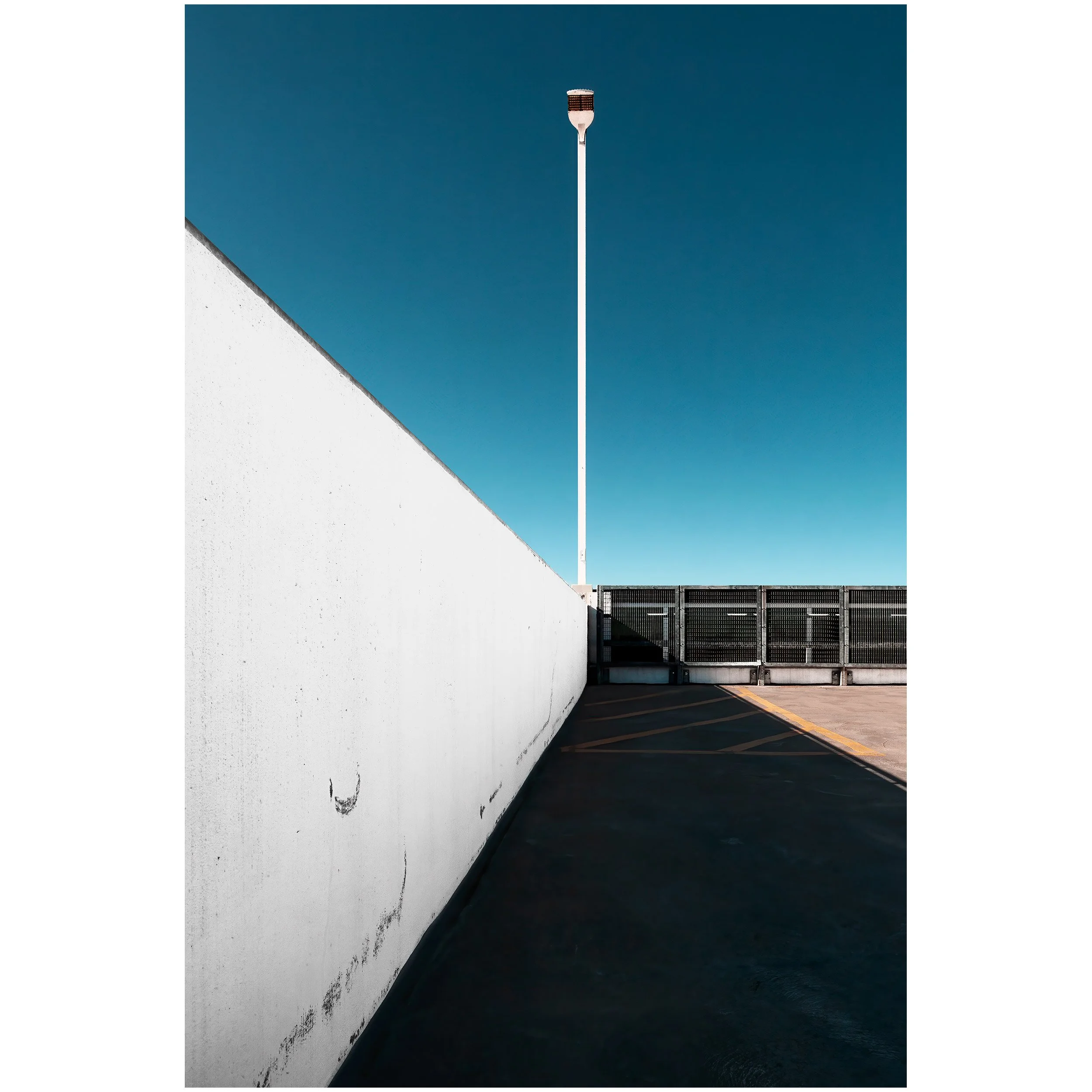 Photo of a blue sky, white wall on the left, and a gated parking lot on the right with a streetlight in the center.