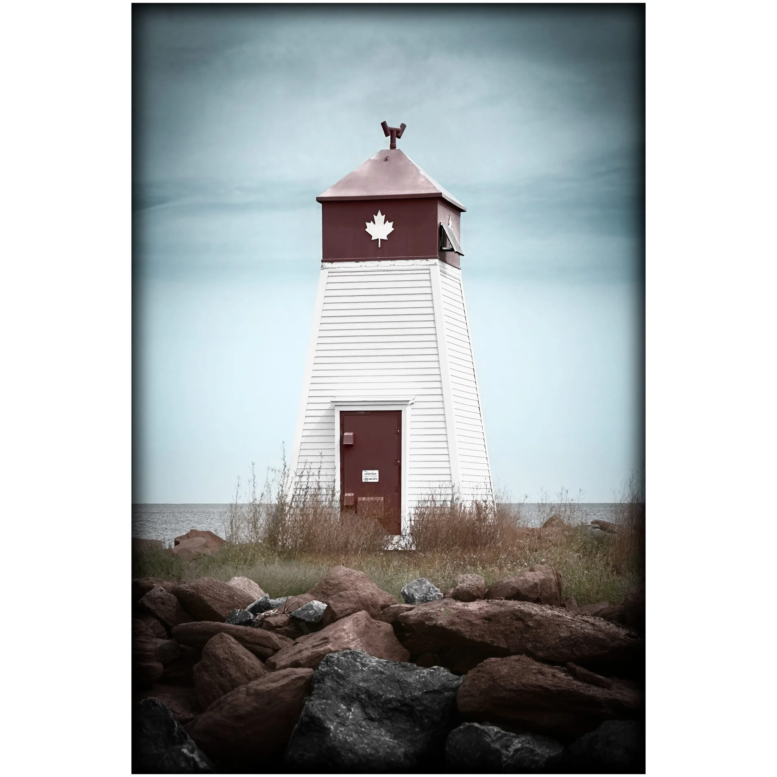 A small white lighthouse with red trim and a maple leaf emblem stands near the water, framed by rocks and dry grass under a pale sky.