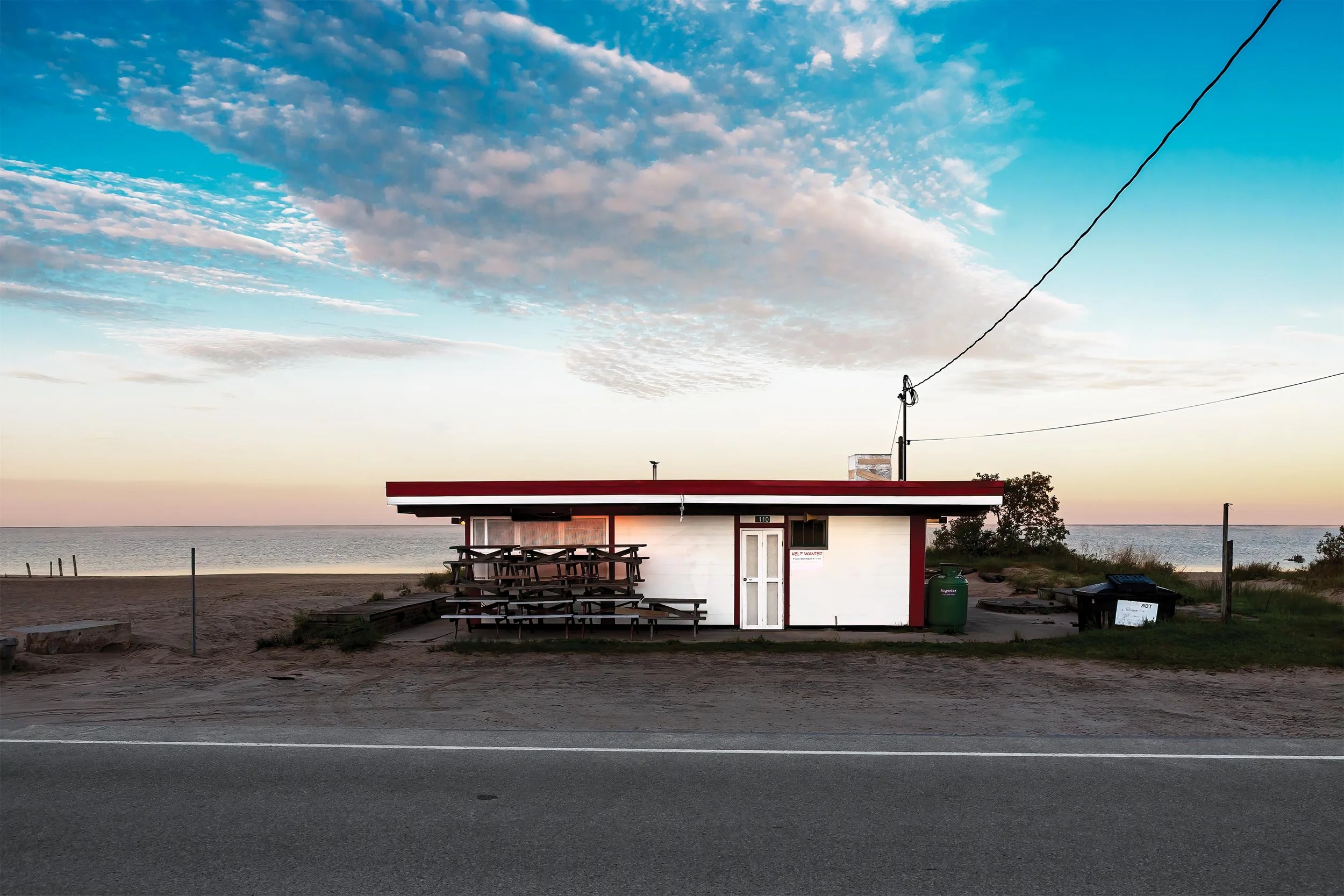 A small building with a red roof and white walls is located near a body of water, with a picnic table and benches outside. The sky has scattered clouds during sunset or sunrise, and there are trees, trash cans, and utility wires visible around the building.