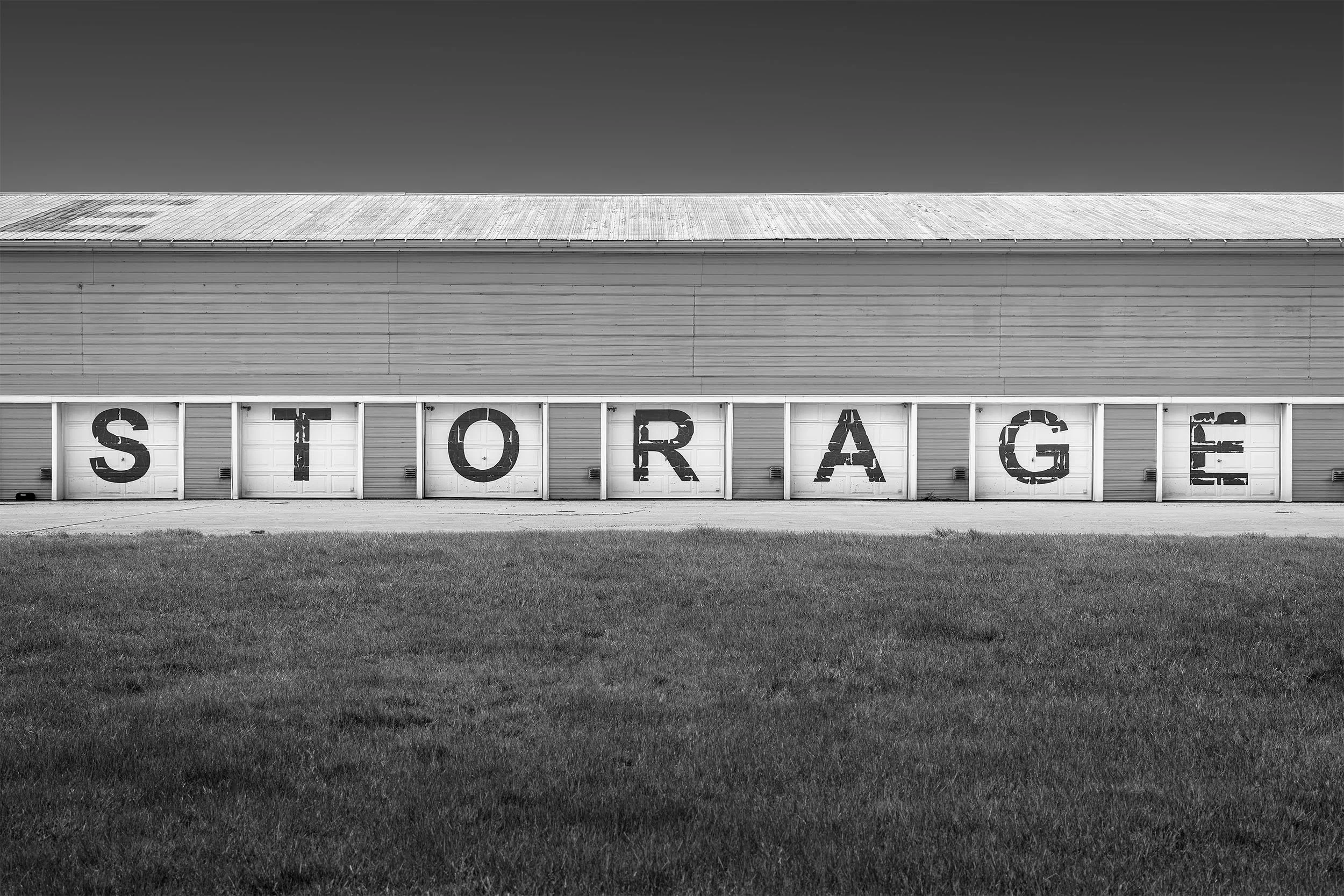 Black and white photo of a large building with garage doors. Each garage door has a letter spelling 'STORAGE'. There is a grassy area in the foreground and a dark clear sky above.